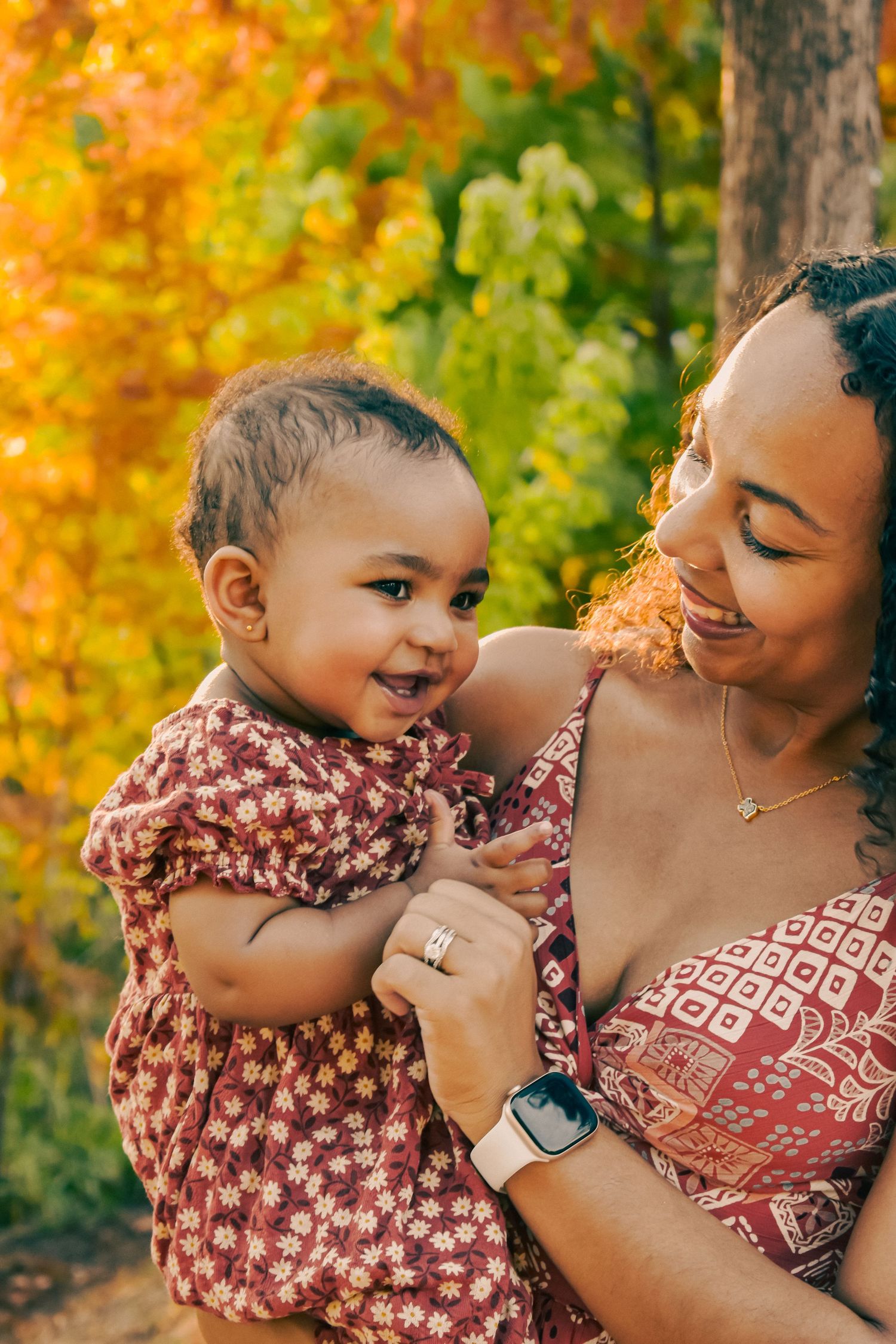 A curly-haired mother smiles warmly at her happy baby girl against a colorful fall foliage backdrop.