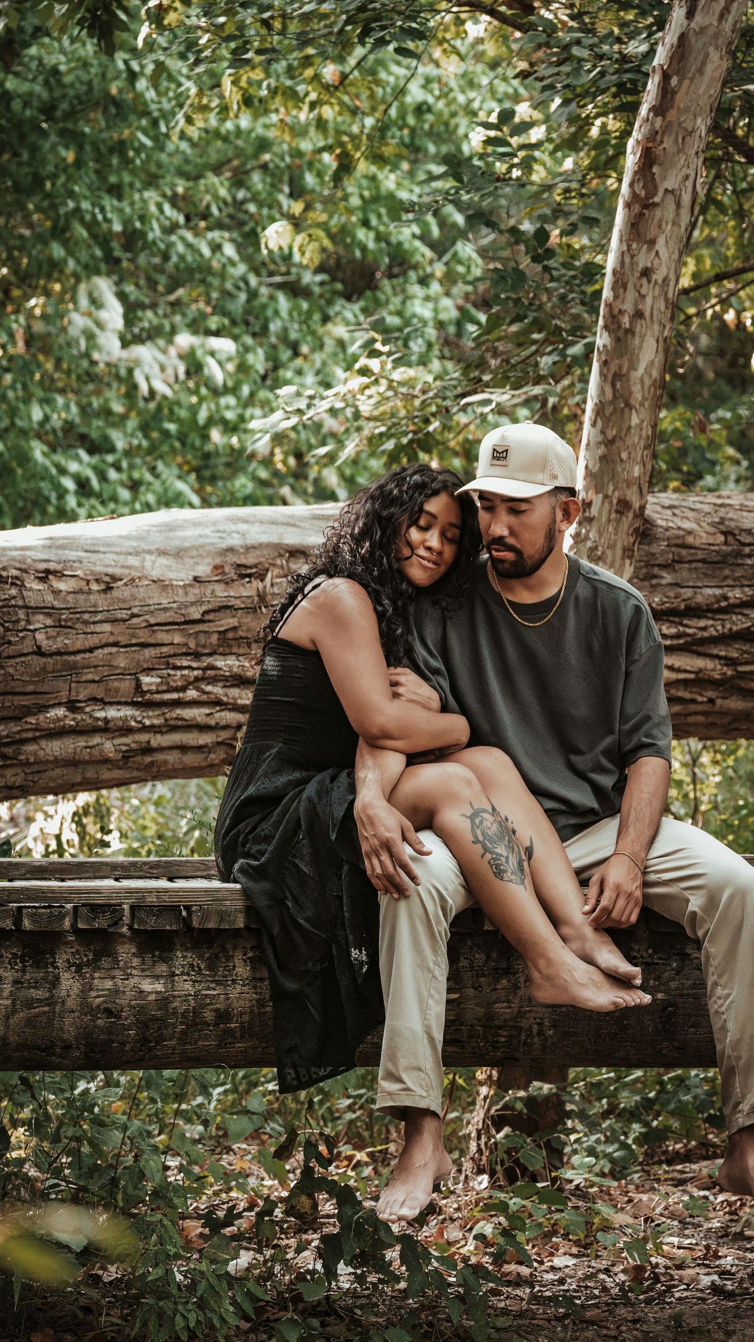 Couple sitting closely together on a fallen log in a green forest, woman leaning on mans shoulder tenderly.