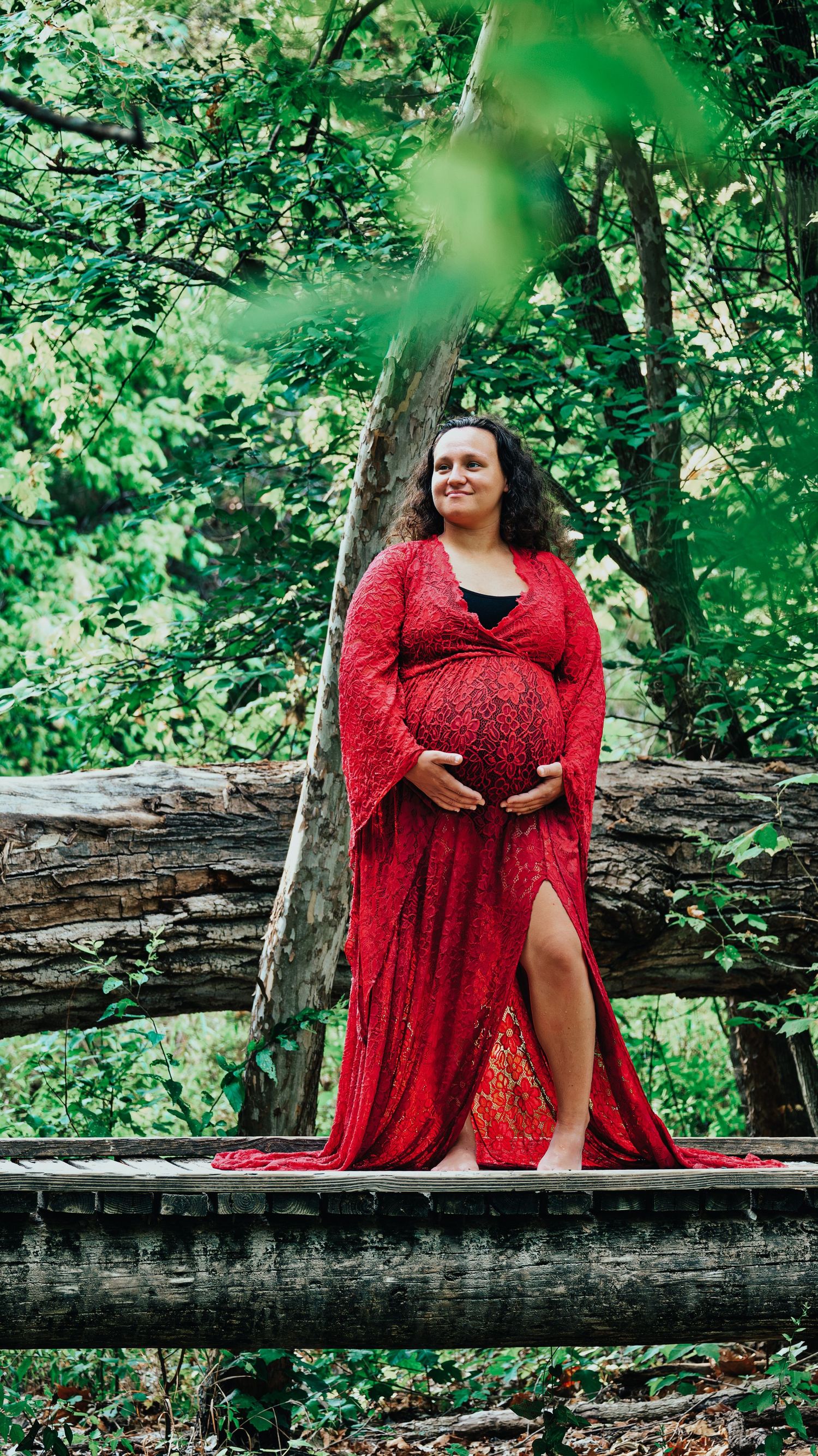 Pregnant woman in elegant red lace maternity dress holds baby bump beside fallen logs in lush green forest.