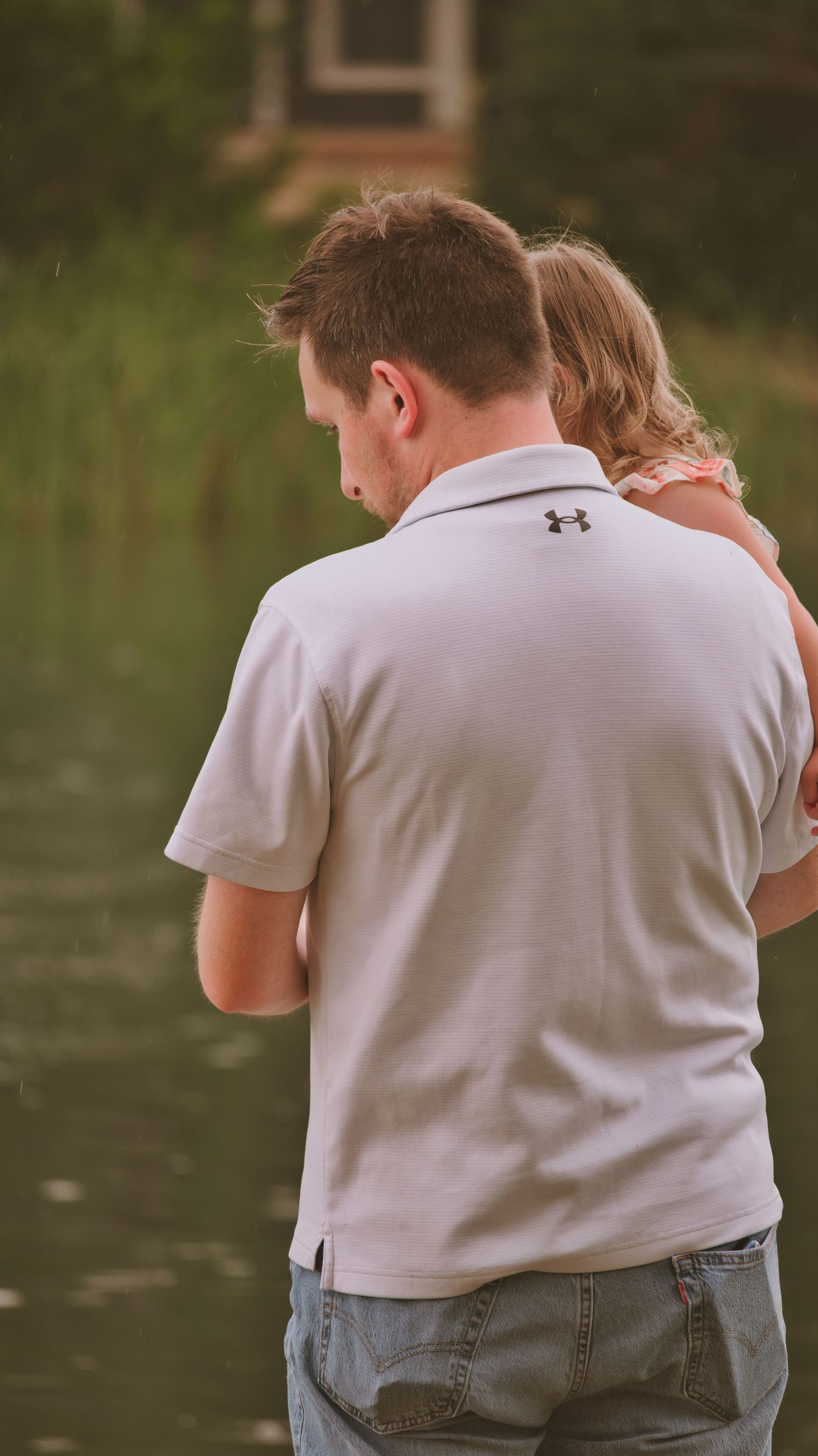 Father in Under Armour polo holds young daughter while standing near a calm pond.