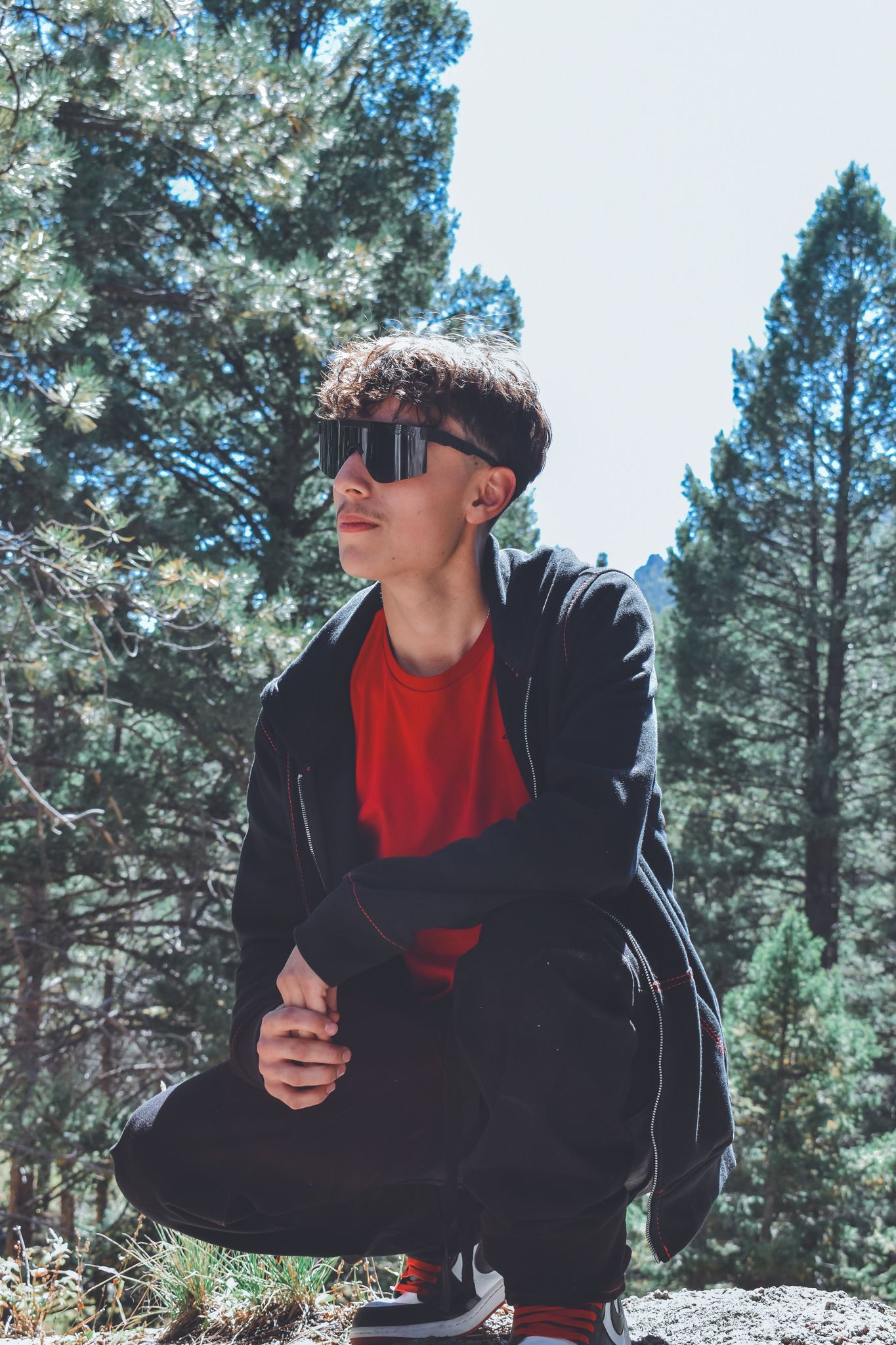 Young man in red shirt black jacket and sunglasses crouching on rock with pine trees behind for senior photos.