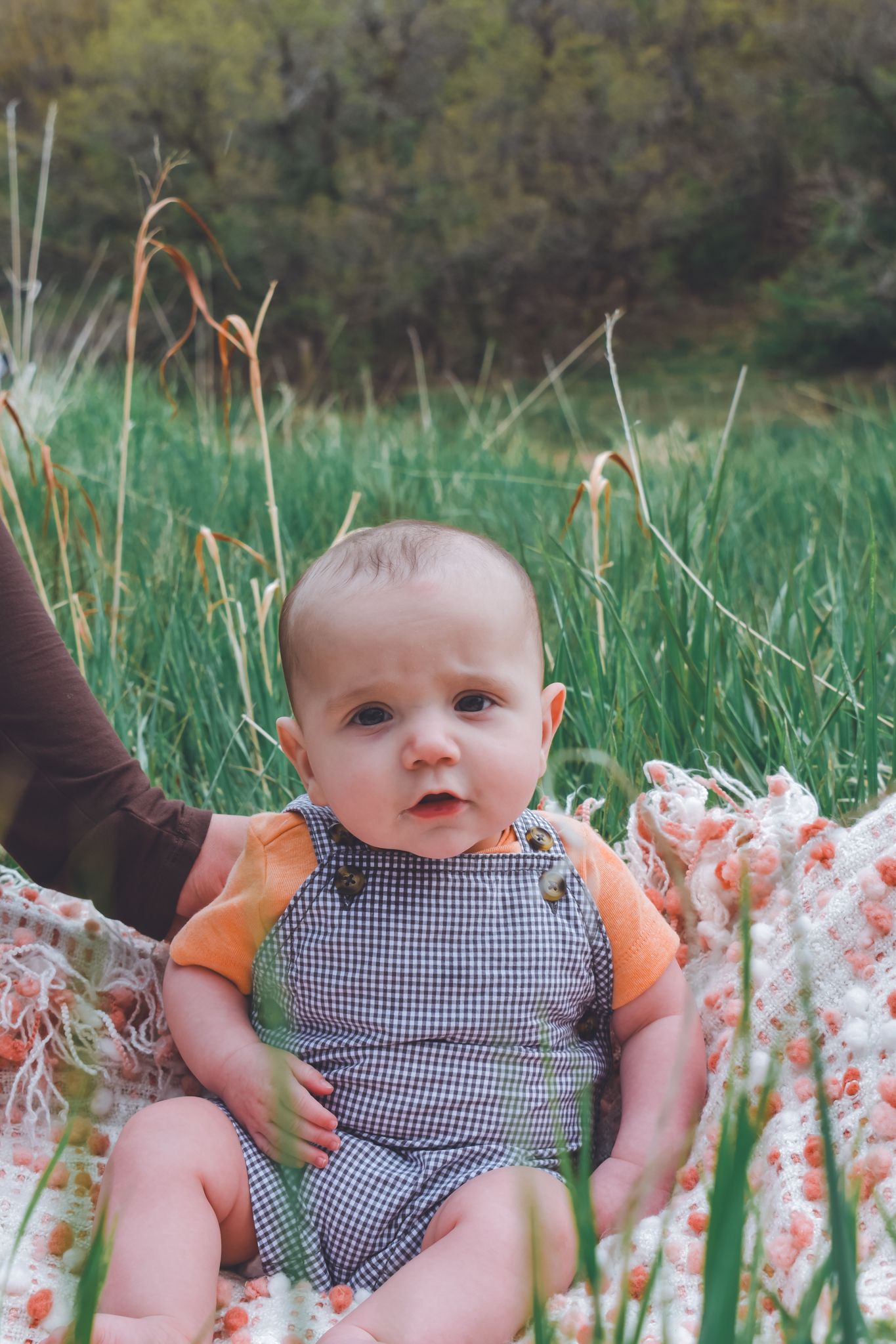 Sweet baby in checkered overalls and orange shirt sitting on floral blanket surrounded by tall green grass.