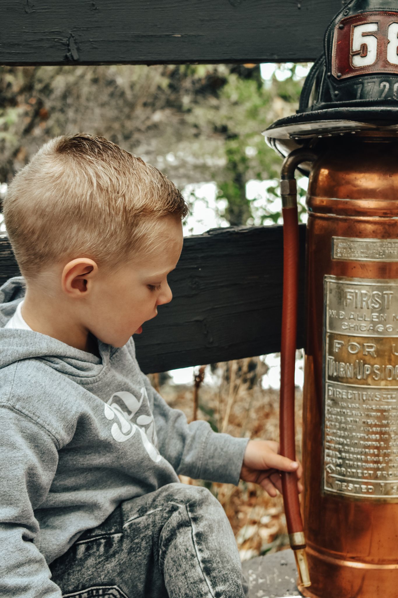 Young boy in gray hoodie examines vintage copper fire extinguisher with number 58 helmet. Family Photographer in Pflugerville, Tx