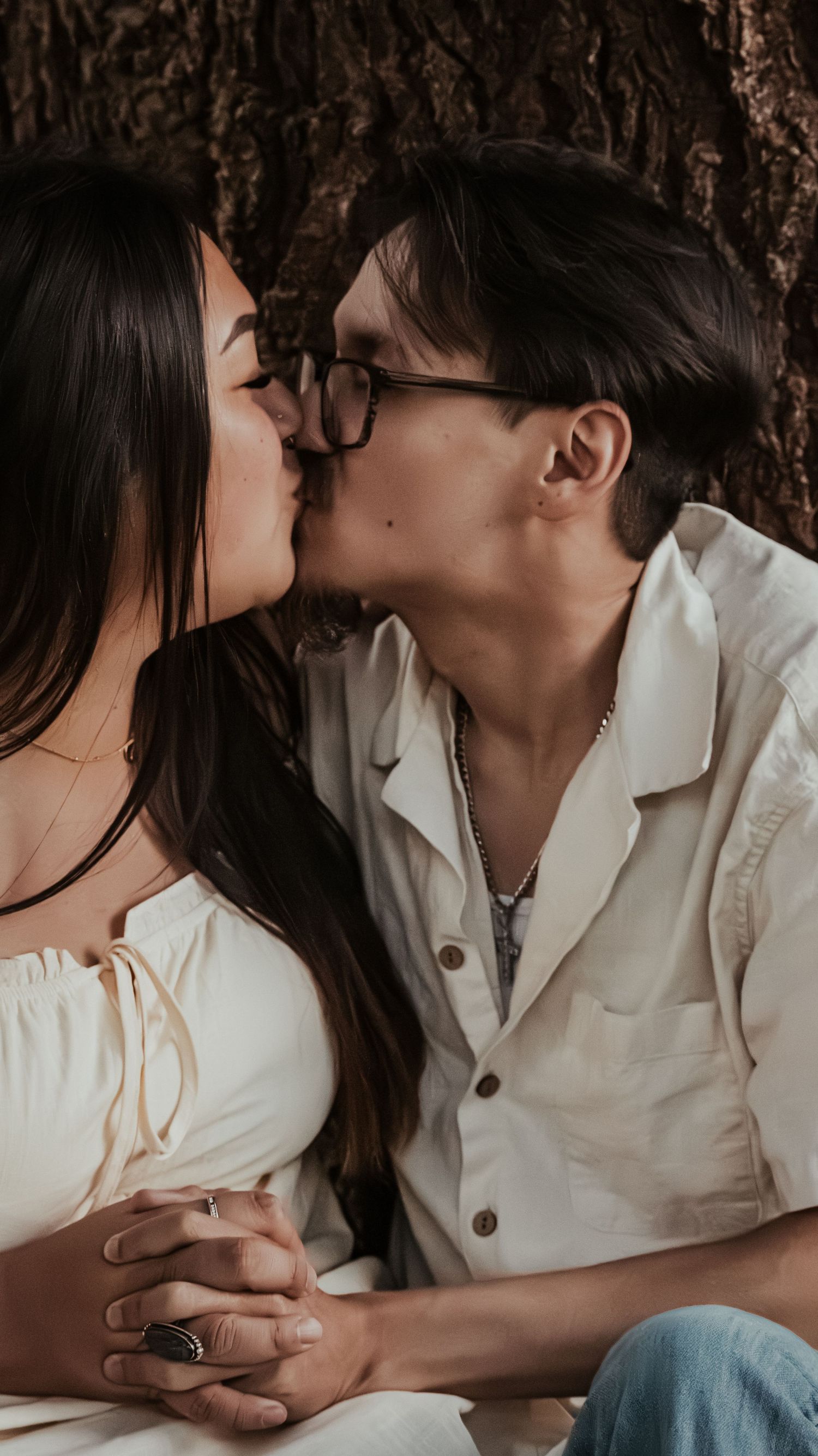 A couple shares a romantic kiss while holding hands seated against a tree trunk.
