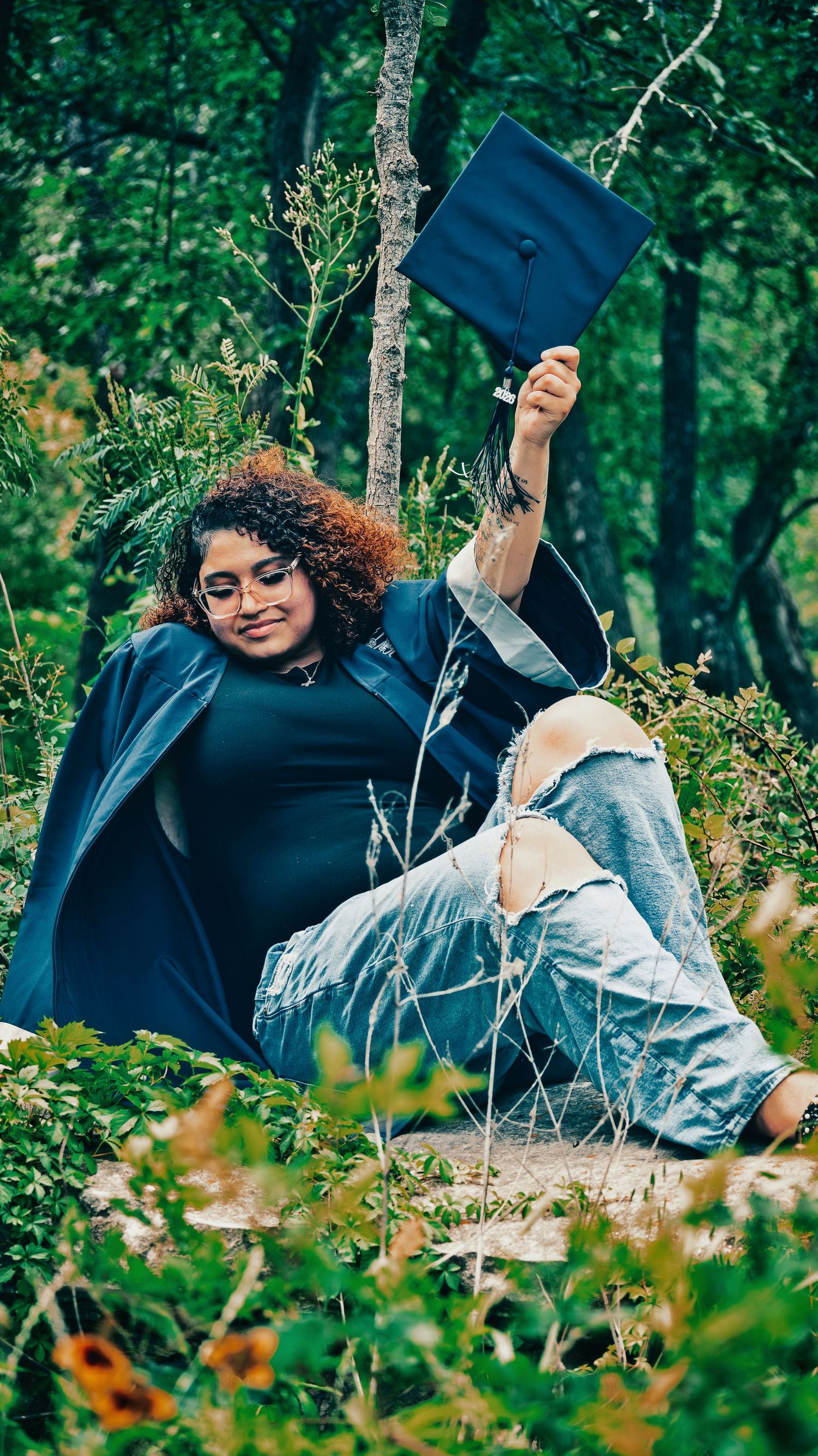 Smiling graduate in blue robe sits outdoors in nature raising mortarboard cap in celebration for senior photos.