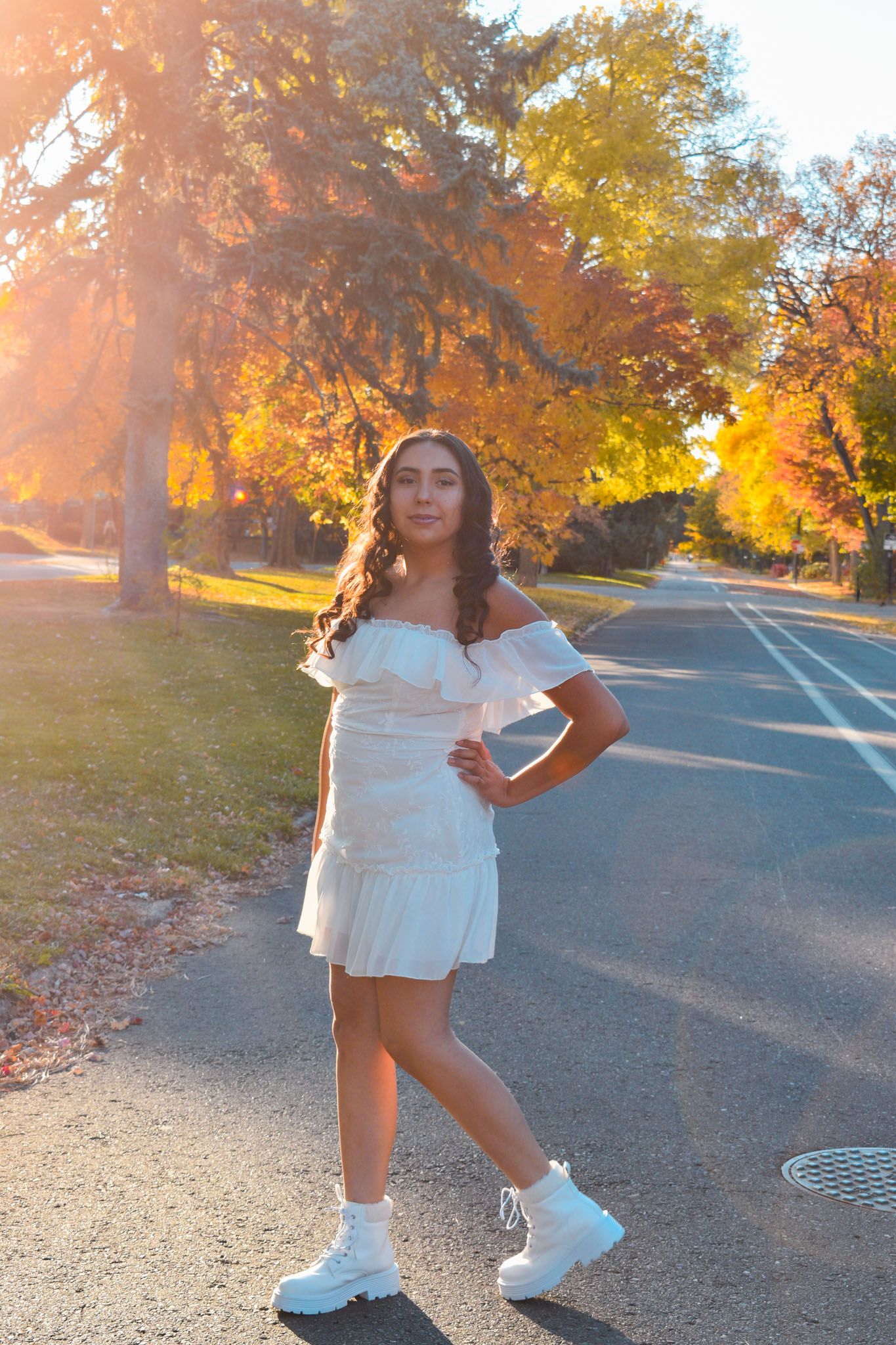 Young woman in white off-shoulder dress posing on a sunlit autumn street for senior photos.