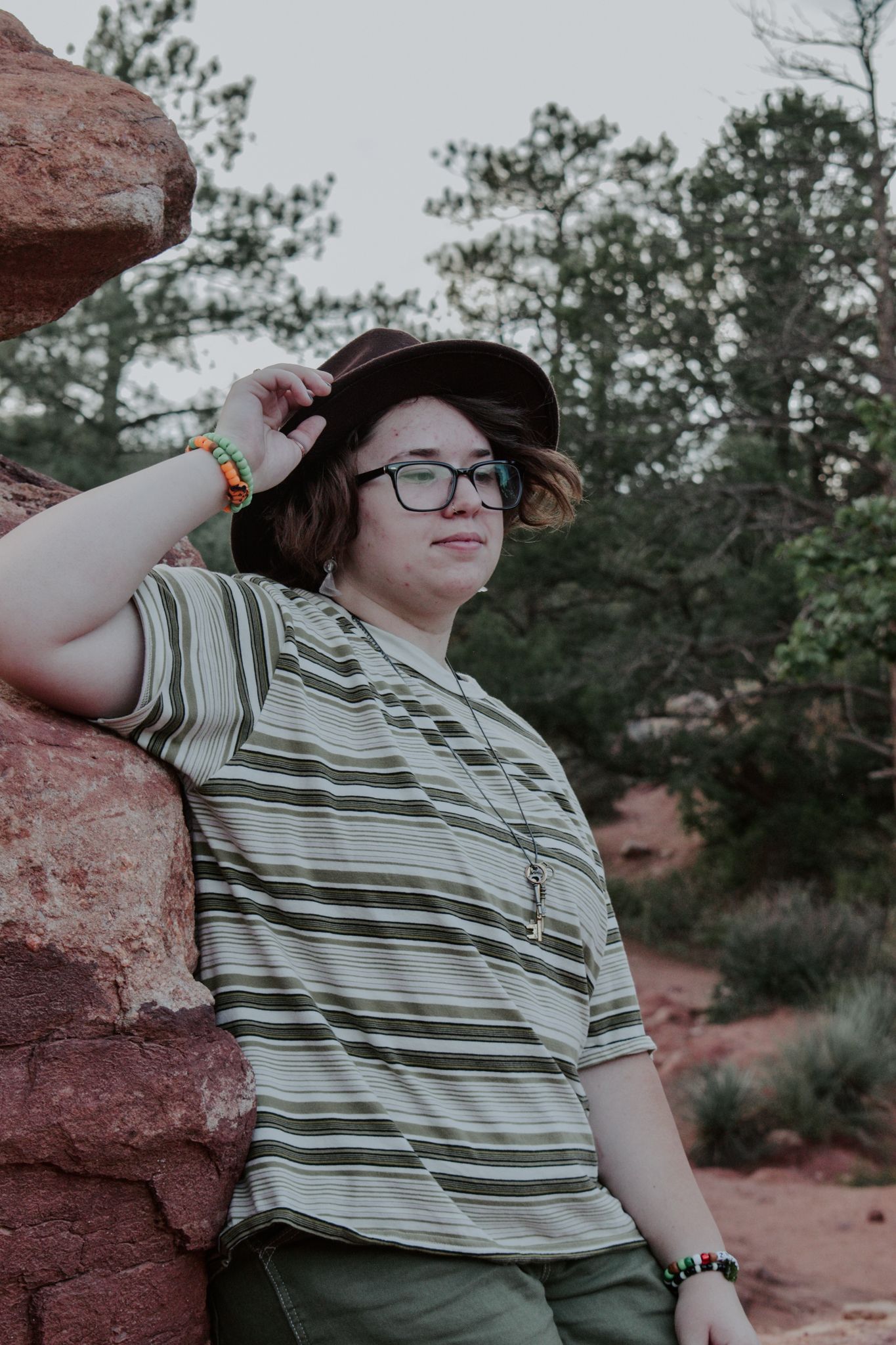 Person leaning against red rock wall, wearing a striped shirt, hat, and colorful bracelet outdoors for senior photos.