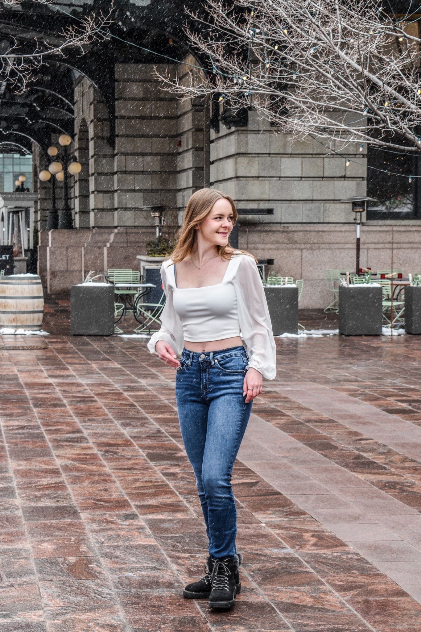 Smiling young woman in white crop top and jeans walking on snowy urban plaza with bare winter trees for senior photos.