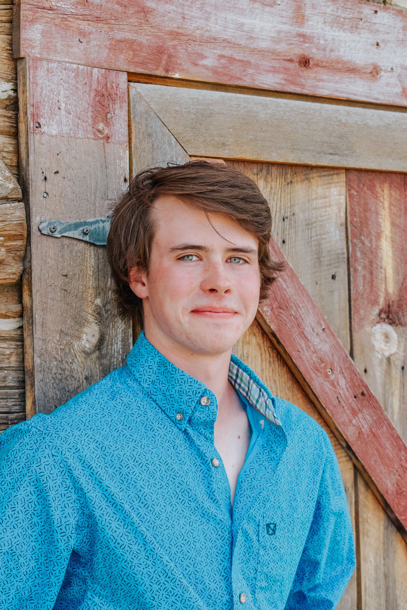 Close-up portrait of young man in teal patterned western shirt leaning against weathered wooden barn wall for senior photos.