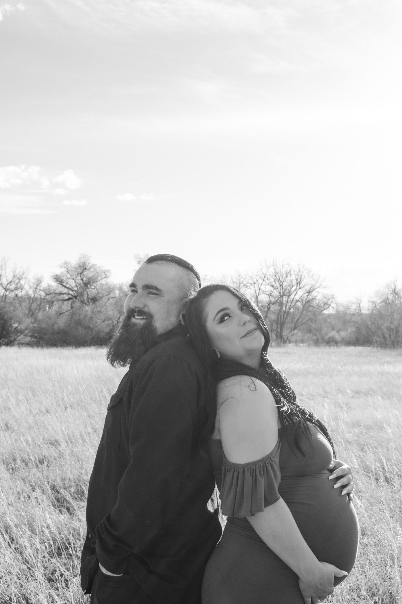 Black and white maternity photo of couple back-to-back in open field under cloudy sky.
