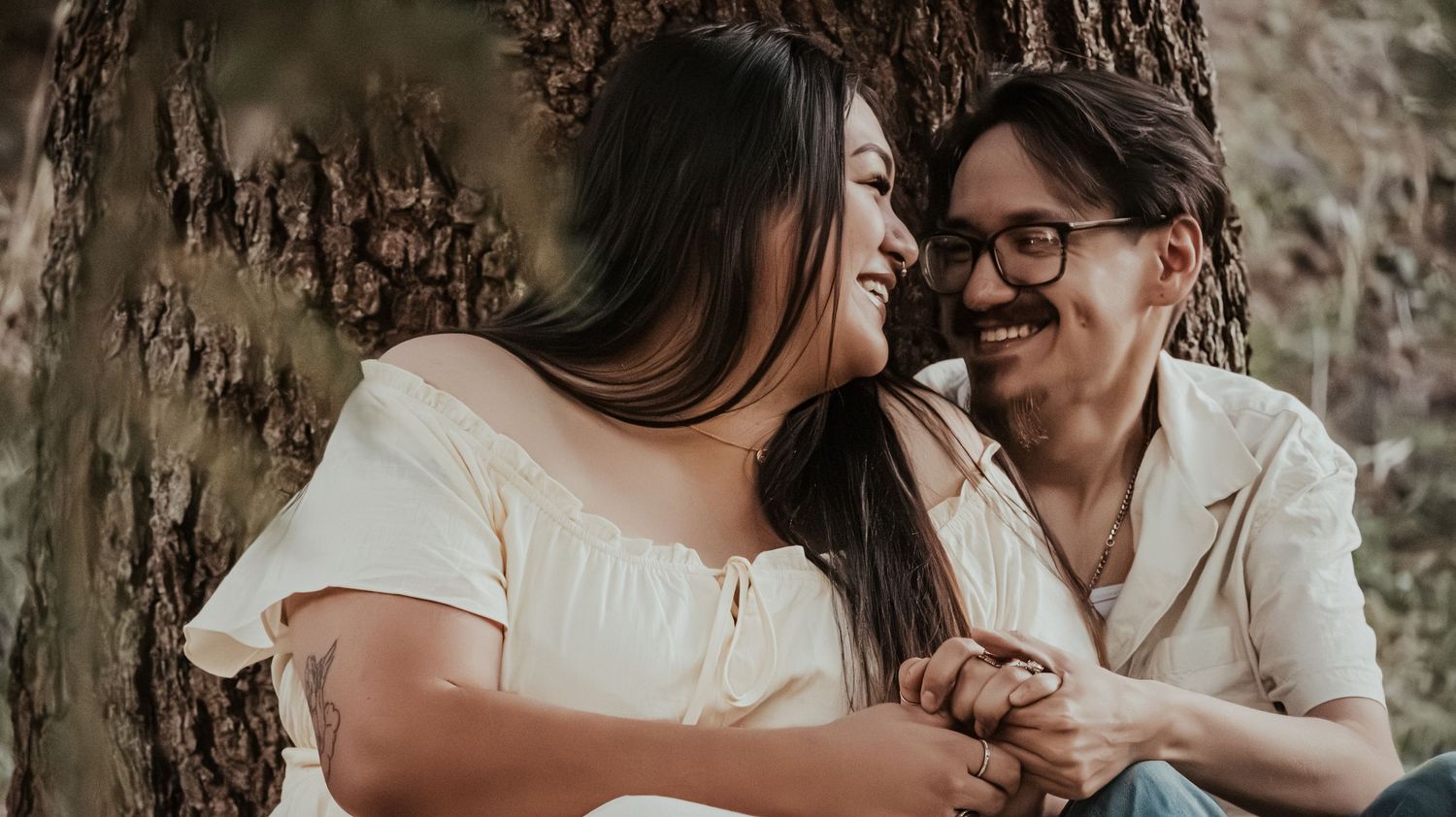 Joyful couple sitting close together holding hands near a large tree.