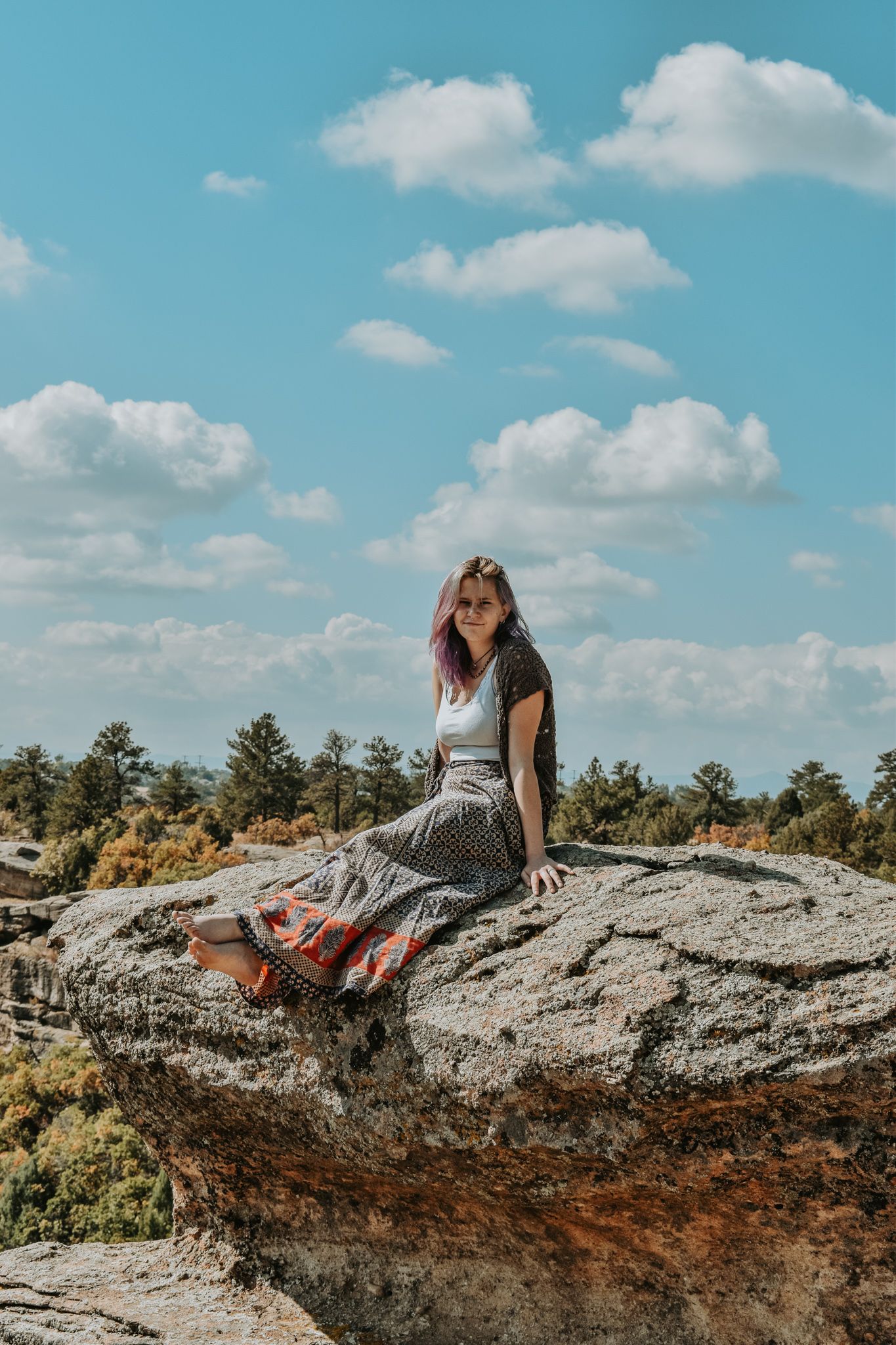 Young woman in boho skirt sits on large rock under blue sky with forest backdrop for senior photos.