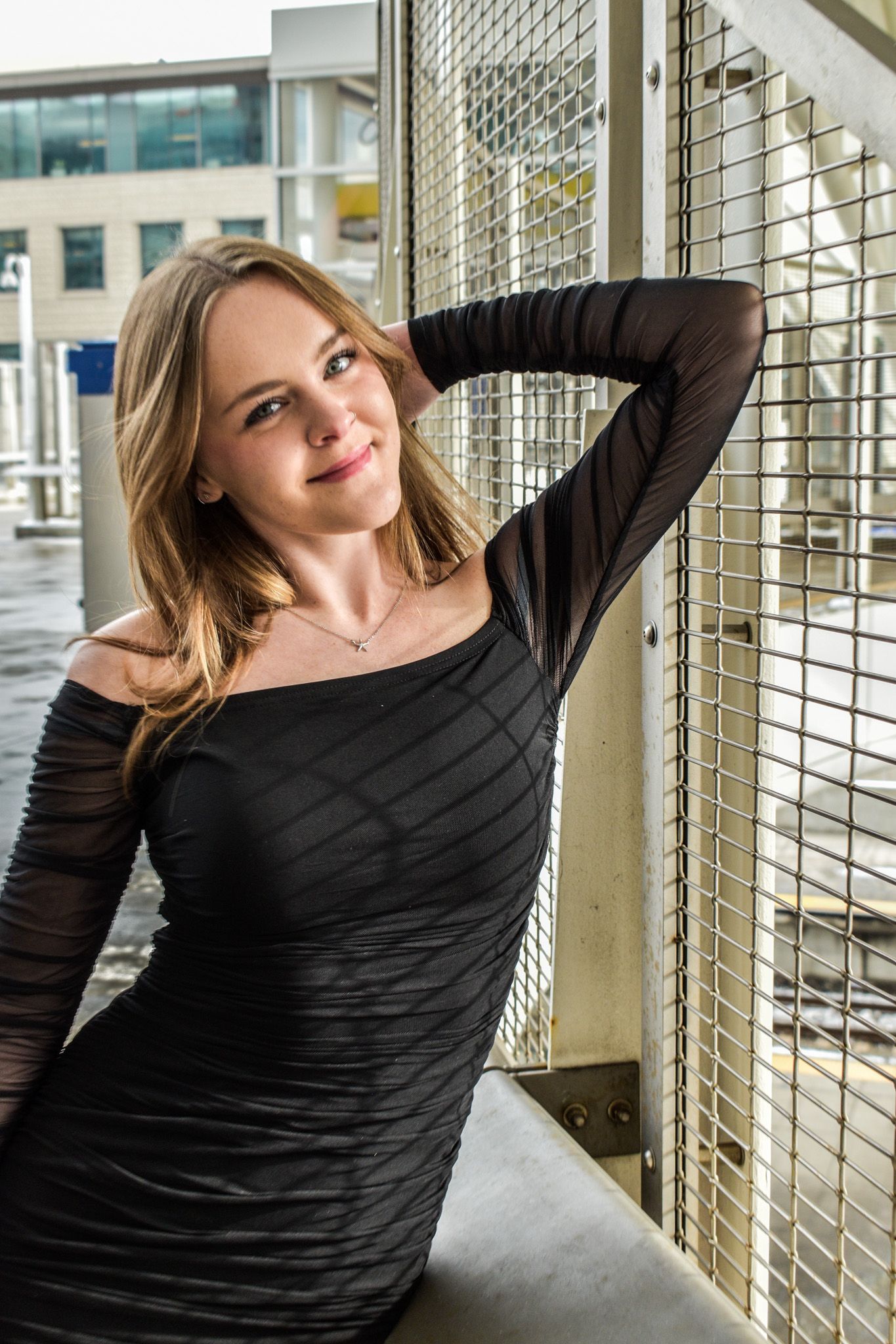 Smiling young woman in black long-sleeve dress posing against metal grid fence at urban location for senior photos.