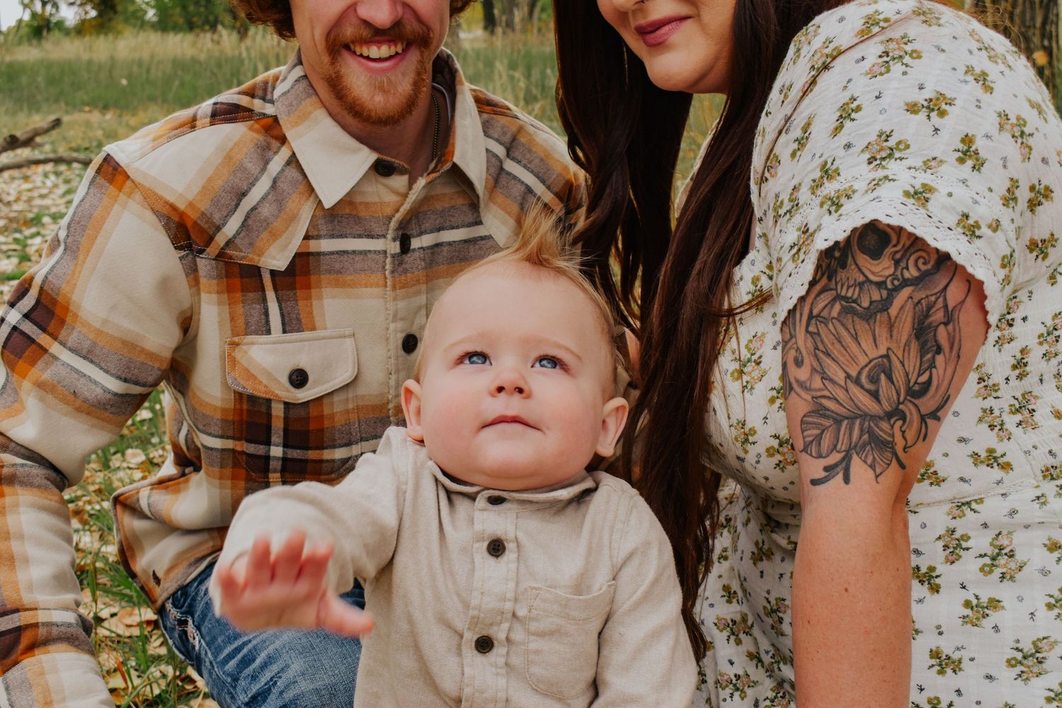 Happy family with tattooed mom and bearded dad sitting outdoors with their baby.