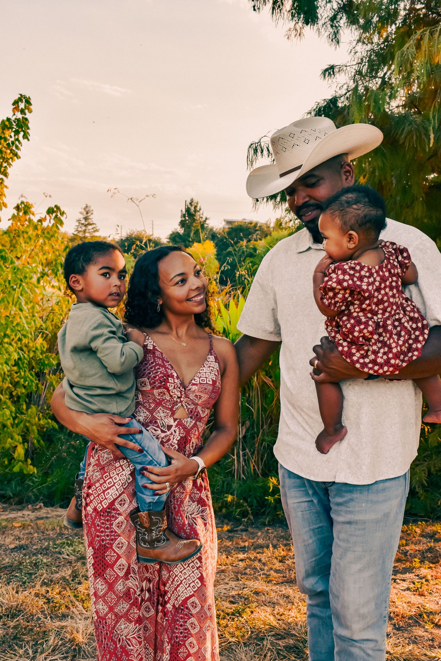 Happy family of four enjoying golden hour outdoors, parents holding two young children near trees.