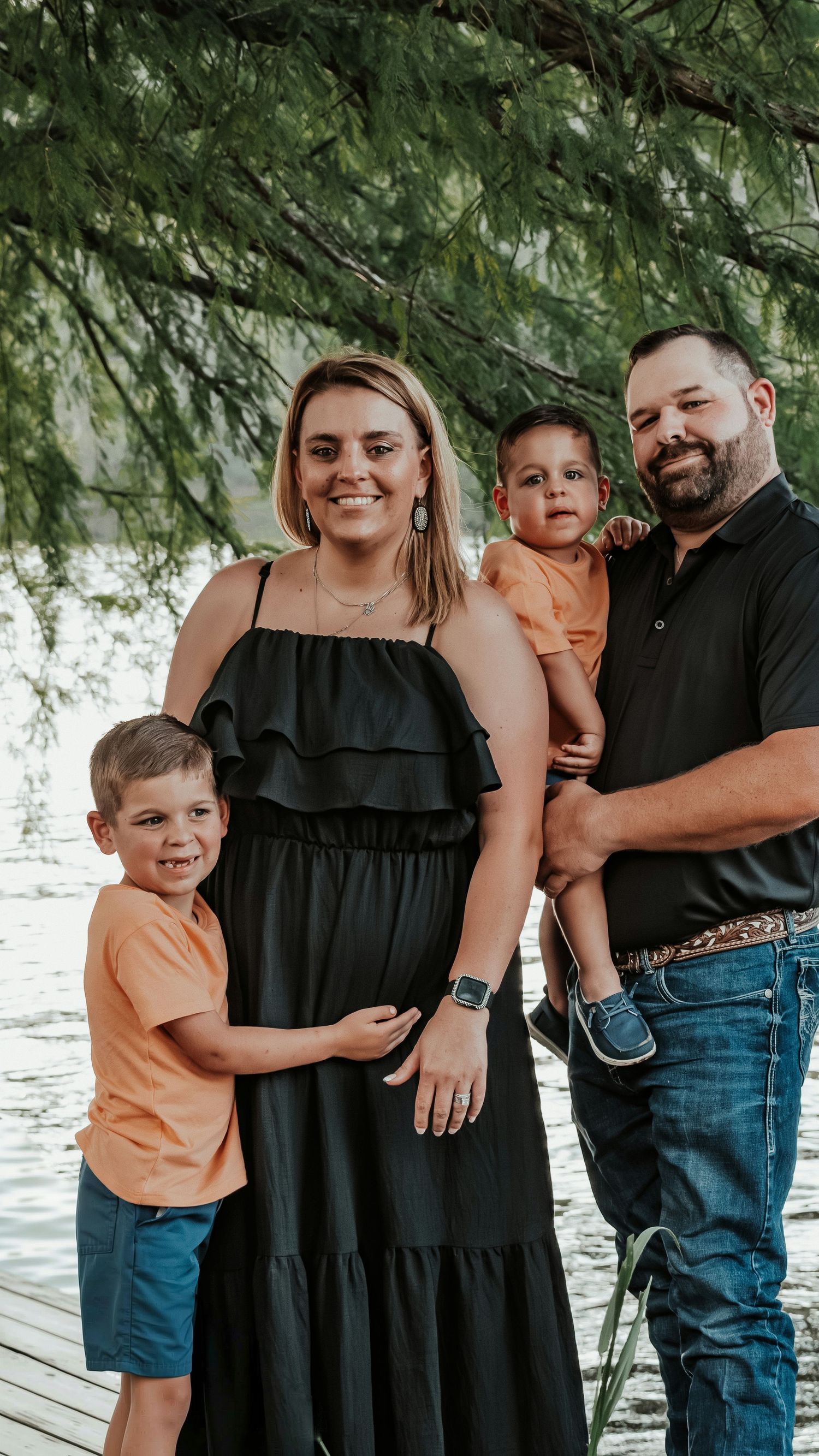 Happy pregnant family of four posing near a lake under green trees.