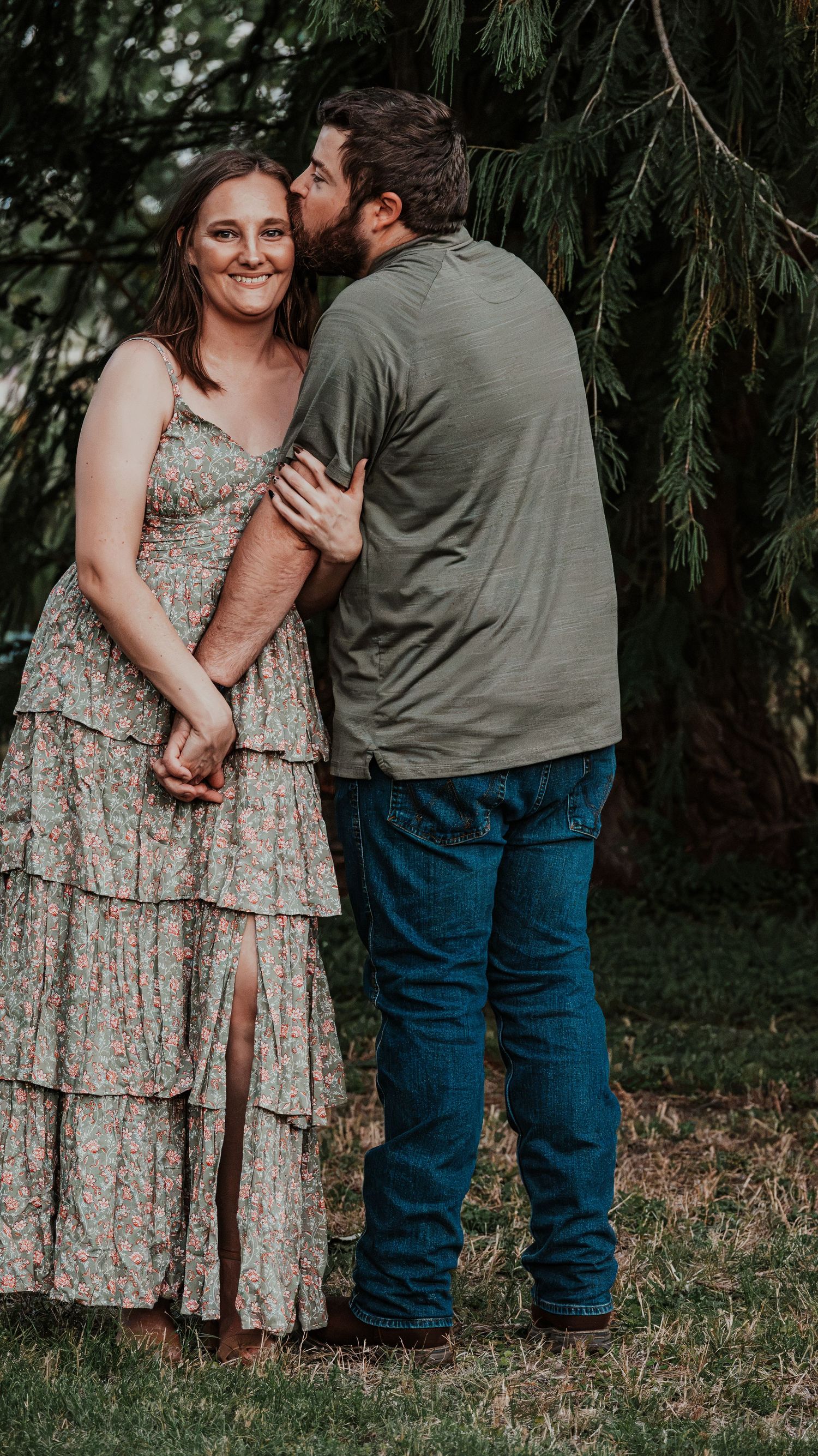 A man kisses a smiling woman in a floral tiered maxi dress while holding her under a pine tree.