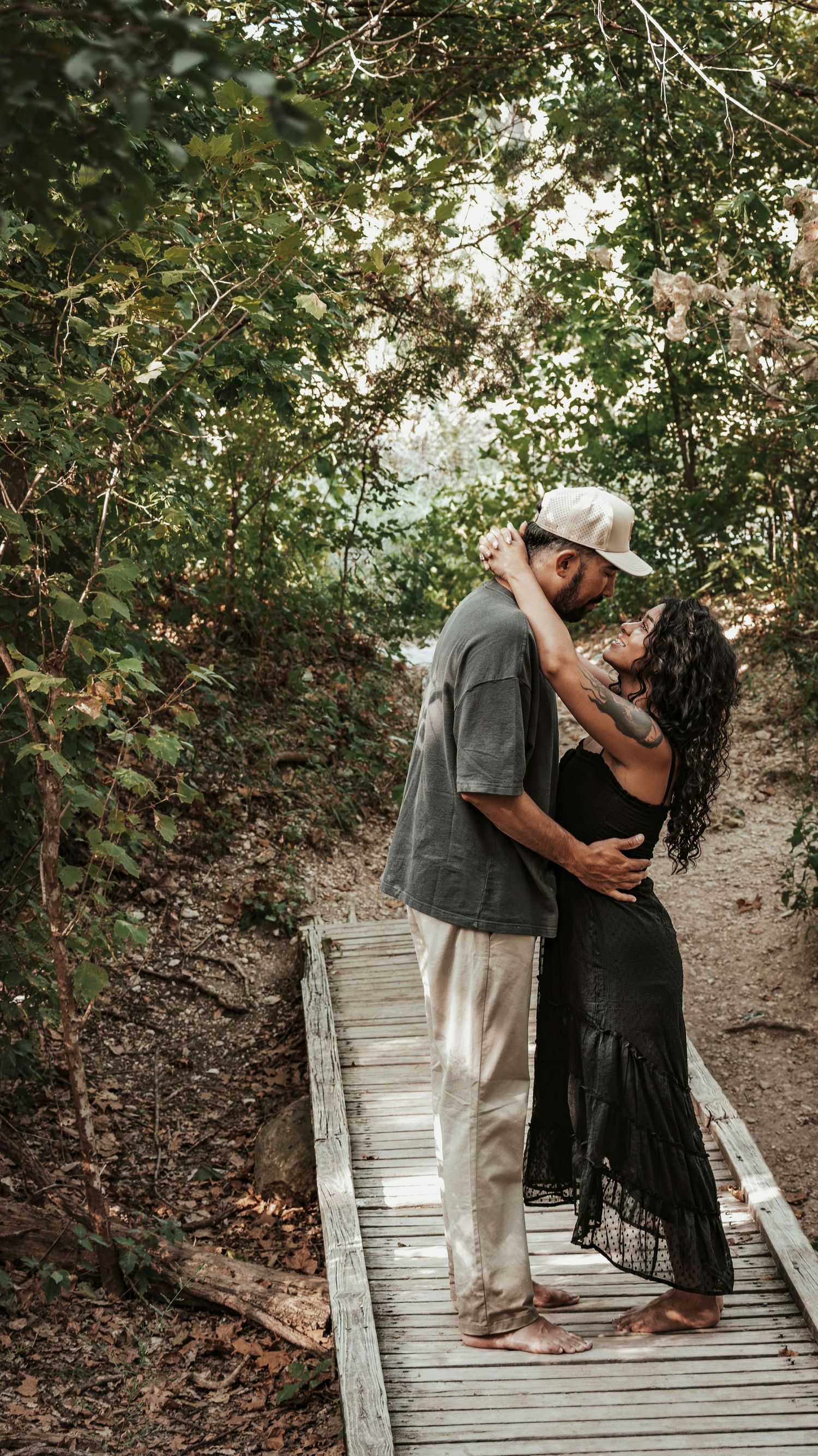 A couple shares a romantic kiss on a rustic wooden boardwalk surrounded by green trees.