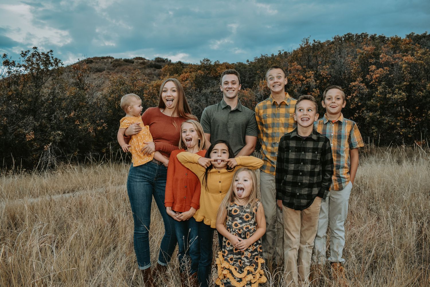 Family of eight children smiling together in a dry autumn meadow with colorful fall foliage behind.