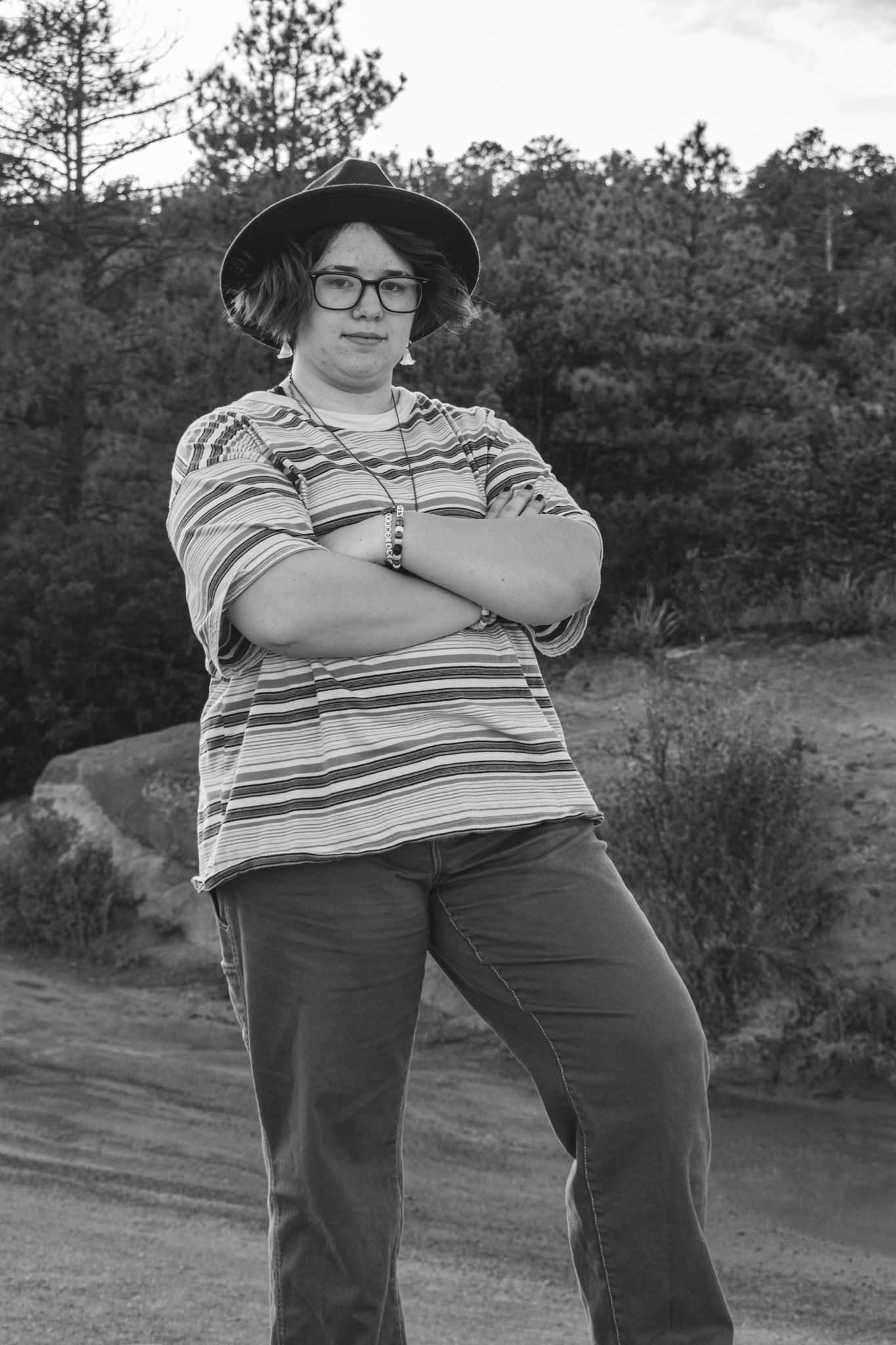 Person standing outdoors with arms crossed in black and white, wearing a striped shirt, hat, and glasses for senior photos.