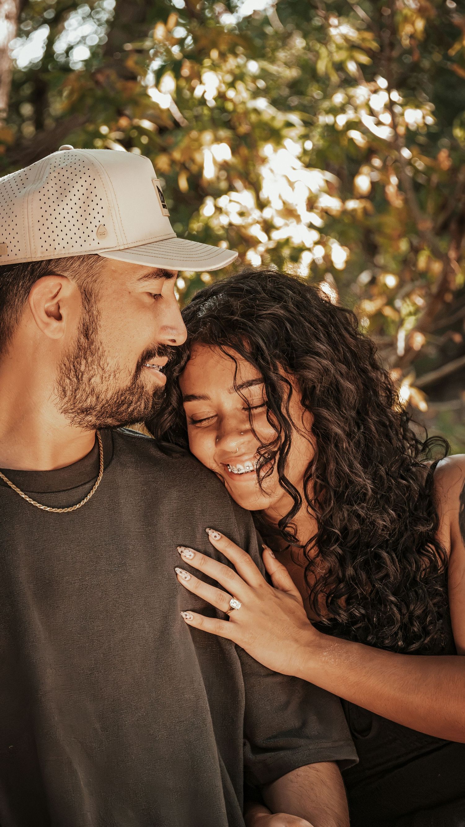A joyful couple shares a tender moment outdoors, the woman displaying an engagement ring for a couples session.