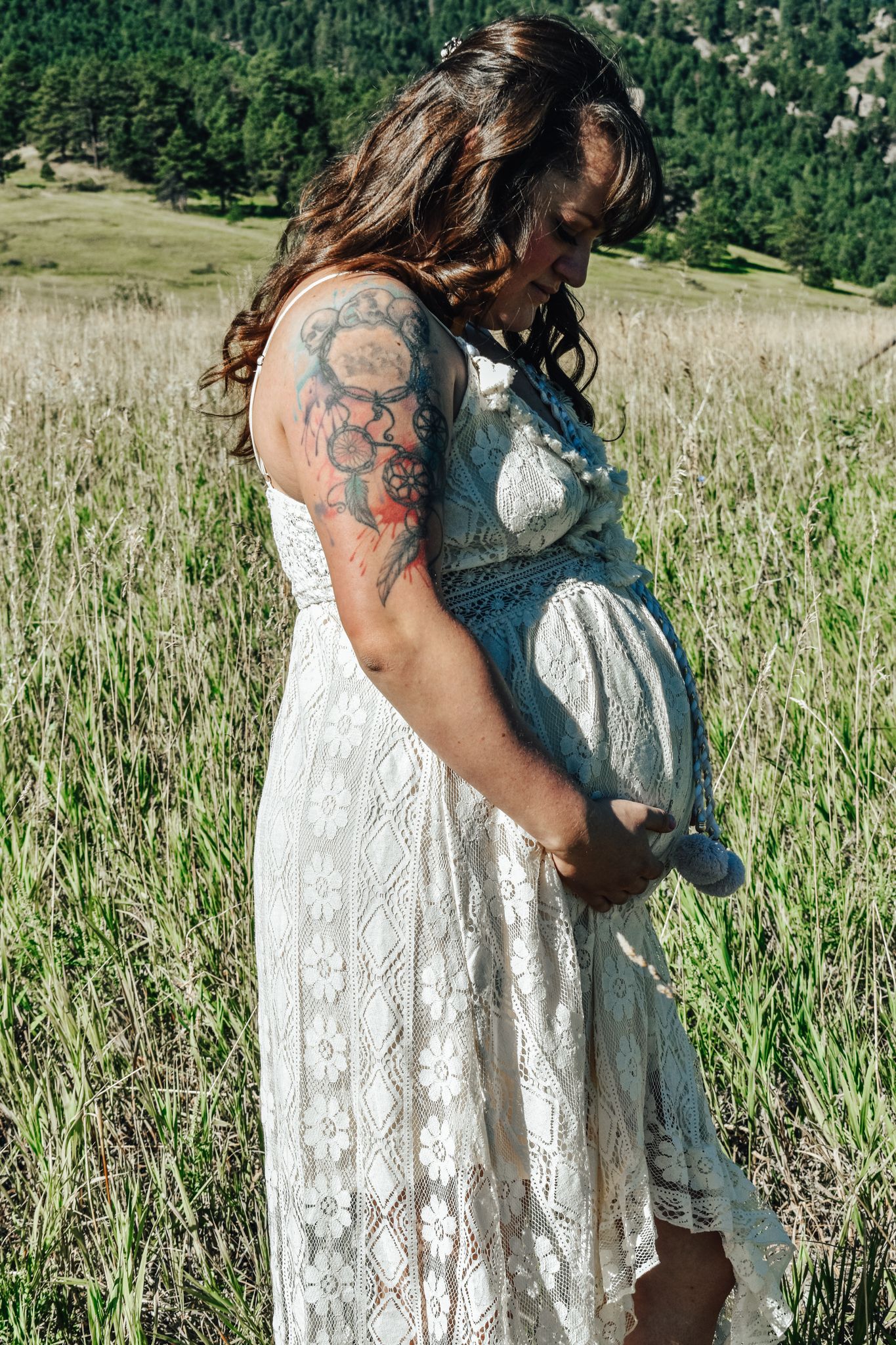A woman in white lace dress with floral arm tattoo standing in green meadow with mountains.