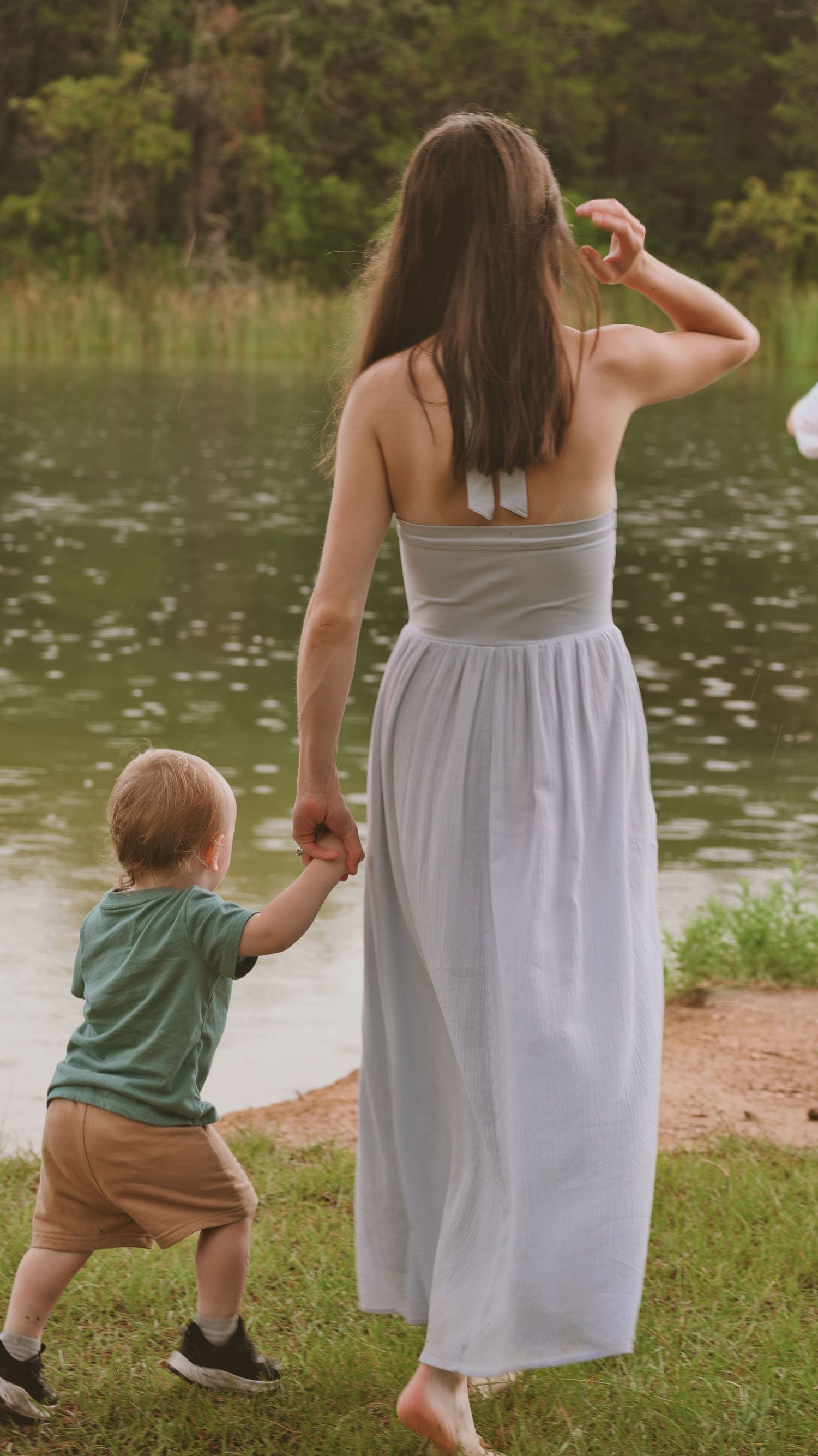 A mother holds hands with her toddler son near a serene lake surrounded by lush greenery for a family session.