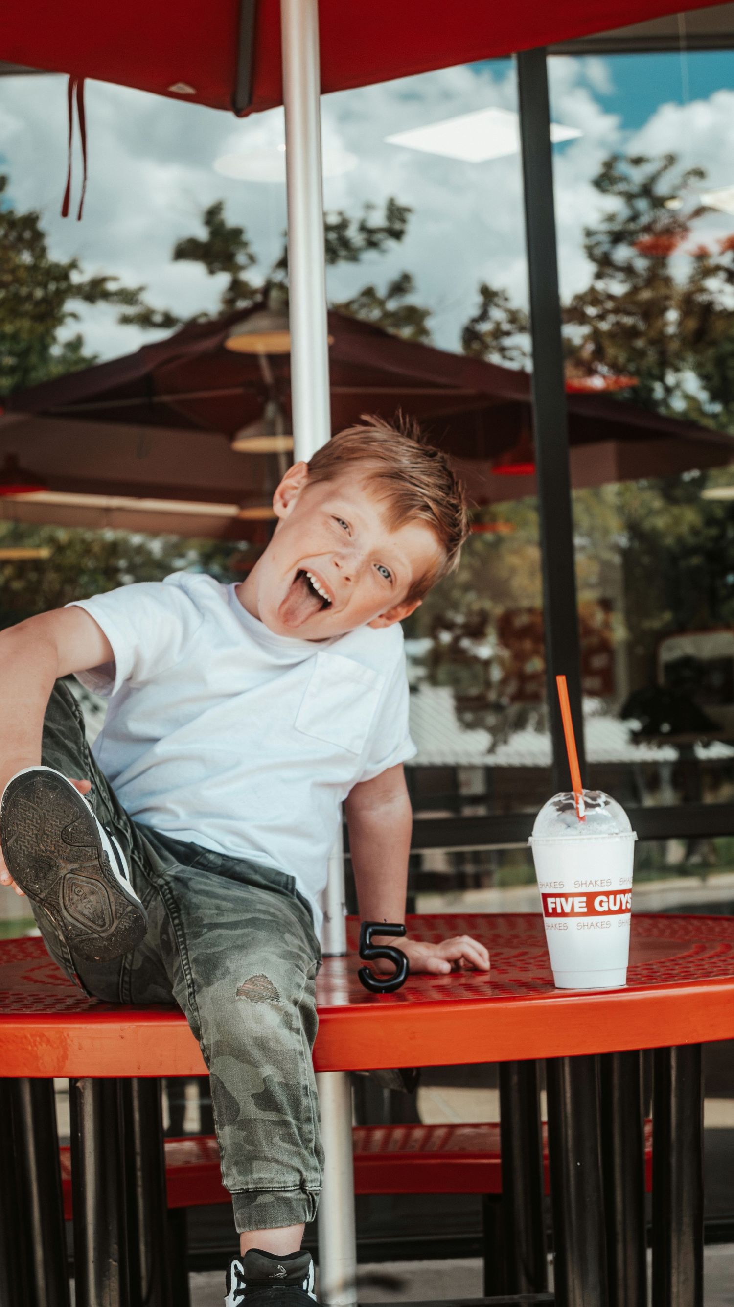 A happy young boy laughing at Five Guys restaurant, sitting on a red table with a milkshake nearby. Family Photographer in Pflugerville, Tx
