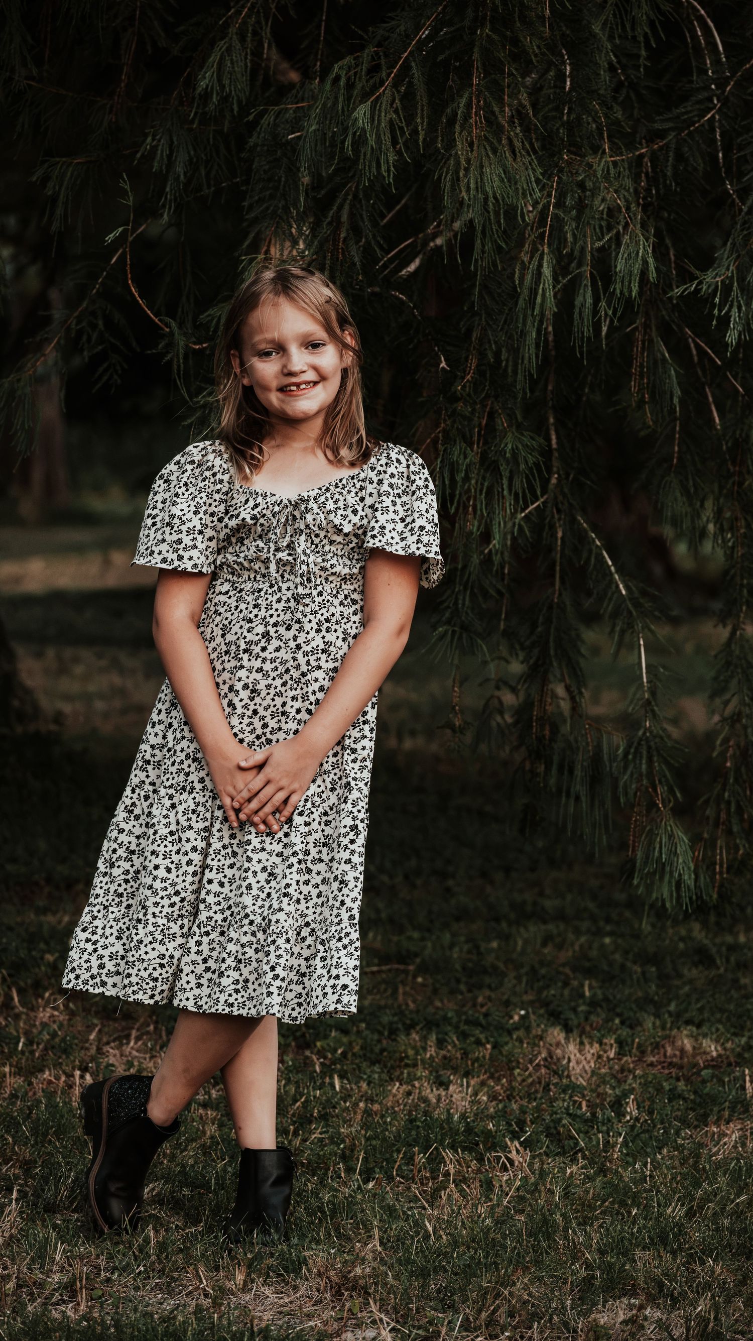 Young girl in floral dress and black boots smiling while standing near pine trees outdoors.