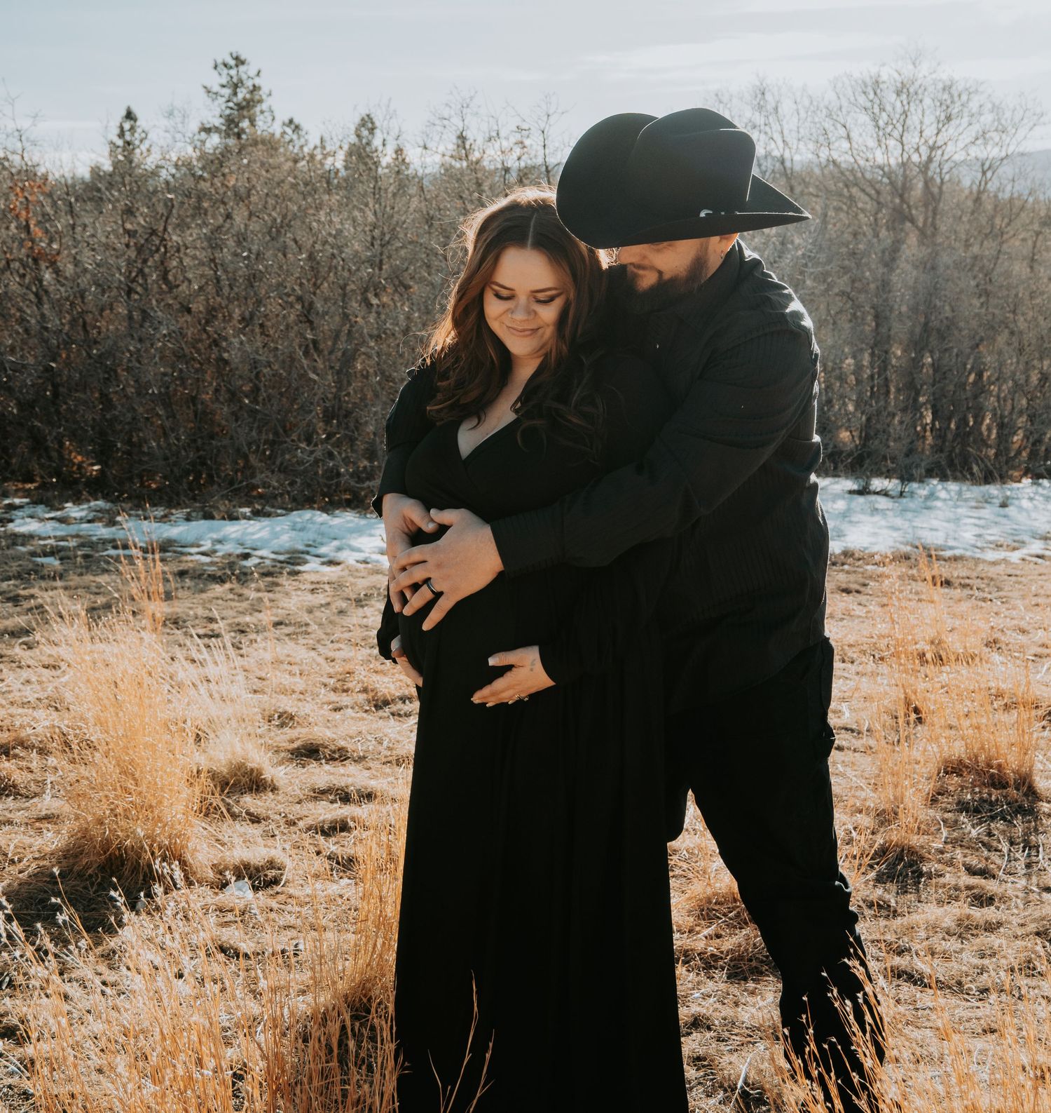 Couple in black embracing outdoors, man in cowboy hat holding pregnant woman's belly in golden field.