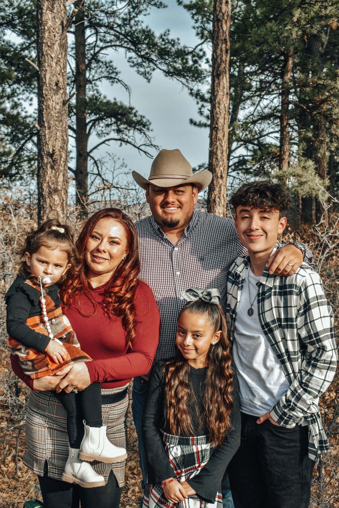 Family of five posing among pine trees with dad wearing a cowboy hat outdoors.