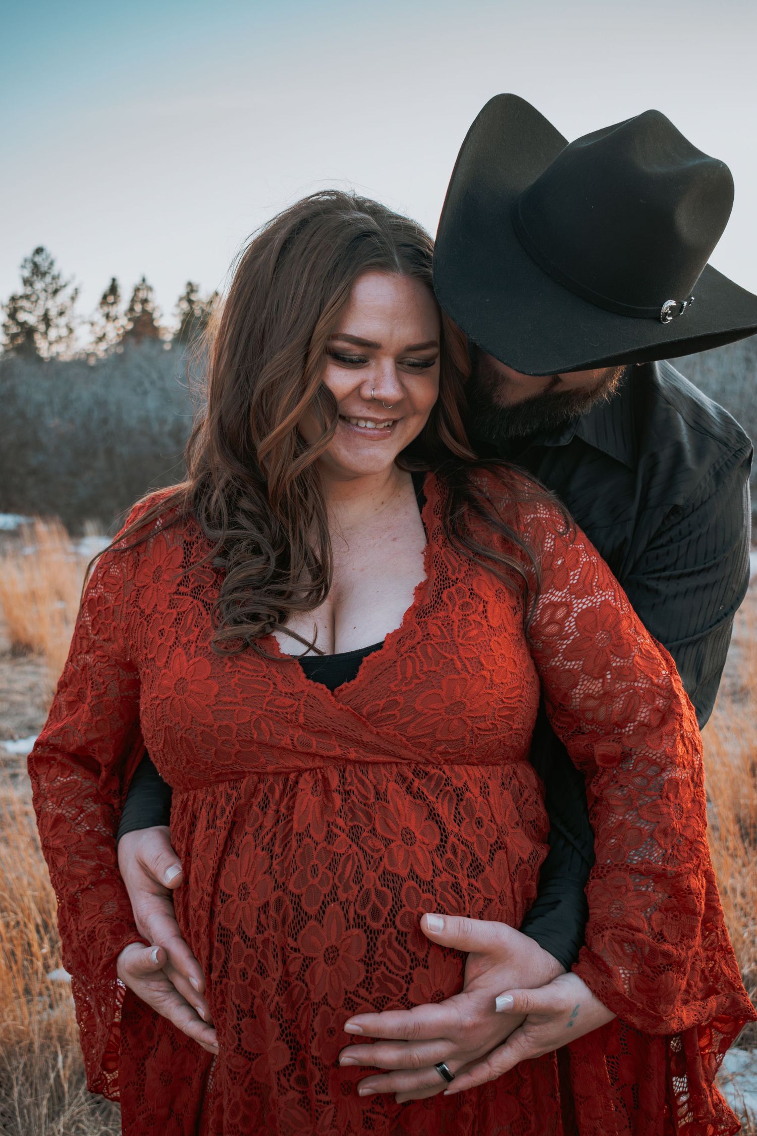Couple sharing tender maternity moment outdoors, woman in red lace dress cradling baby bump while man in black cowboy hat embraces her.