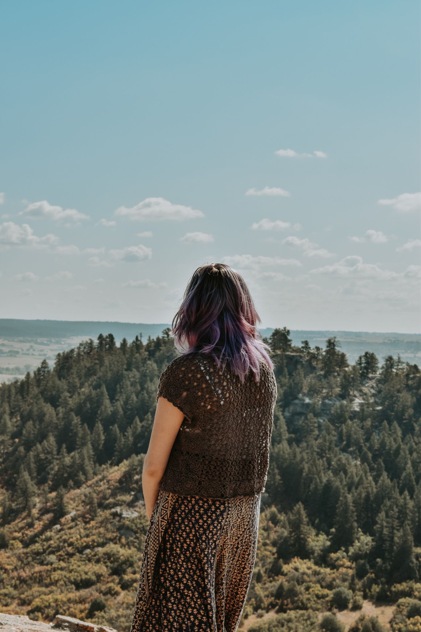 Woman with purple hair stands on rocky overlook gazing at forested landscape for senior photos.