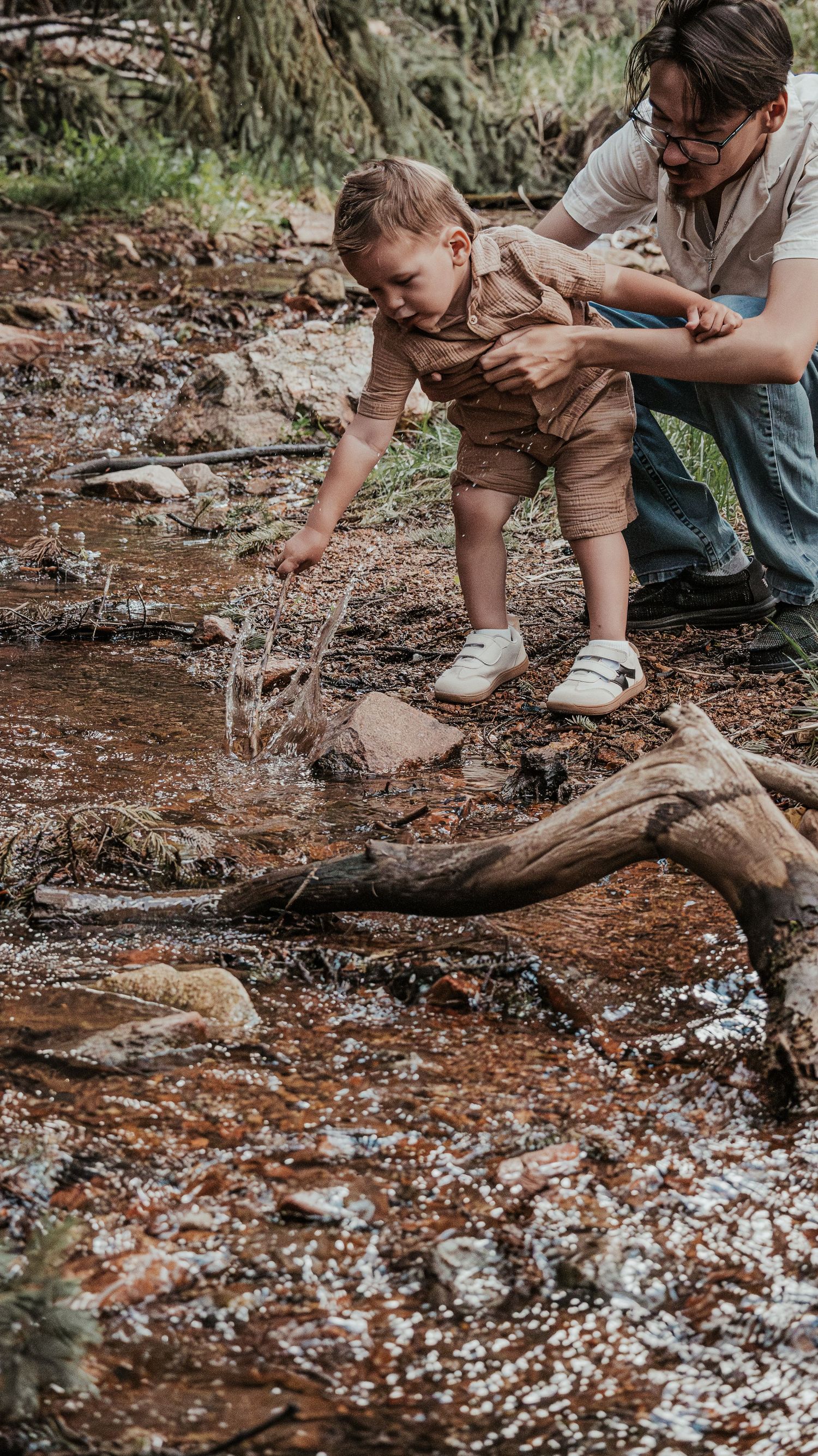 Father helping young toddler boy splash water in a rocky forest stream during family outing.