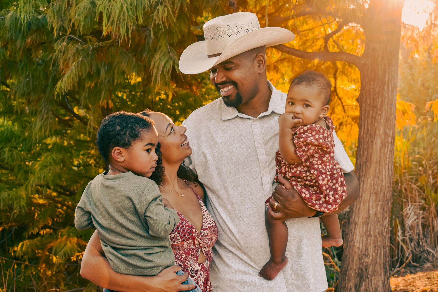Smiling father in cowboy hat holding two young children while mother looks up lovingly outdoors.