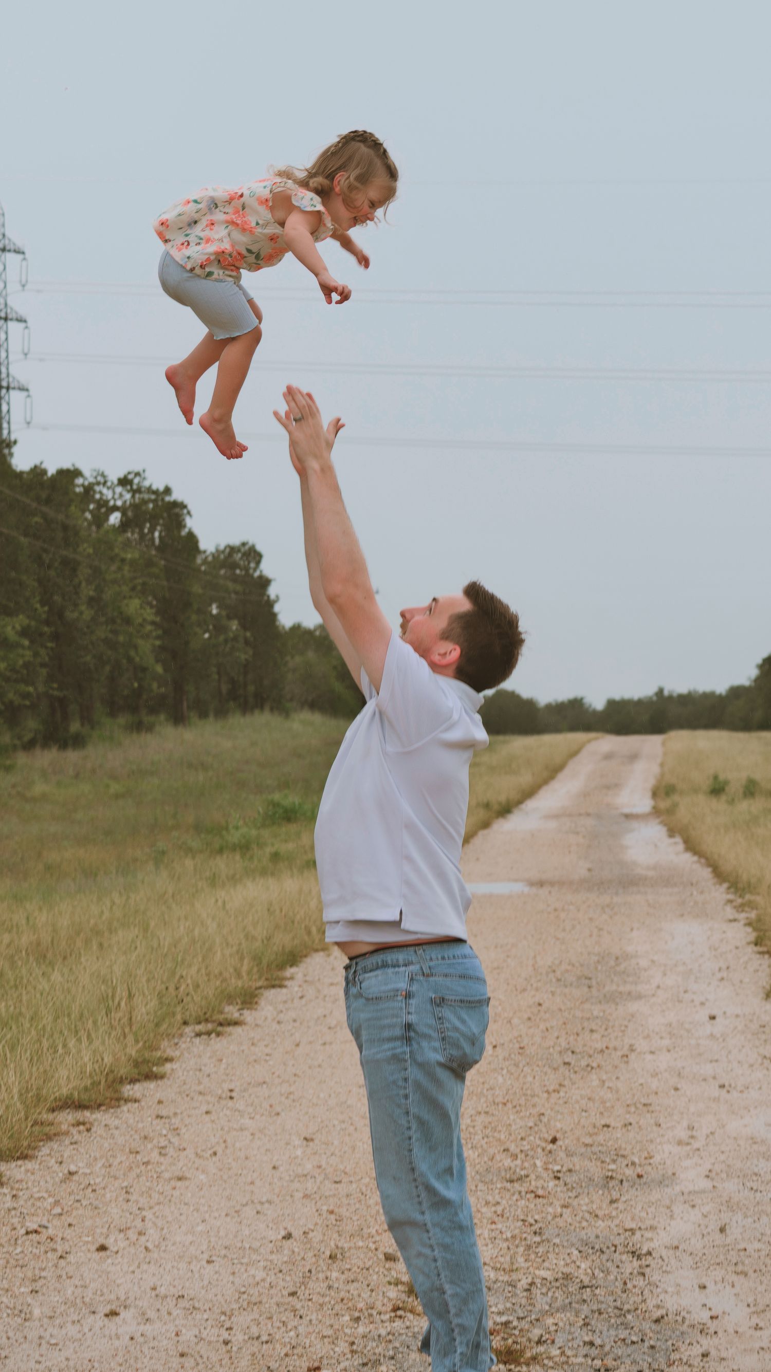 A father joyfully tosses his young daughter high into the air on a rural dirt road for a family session.