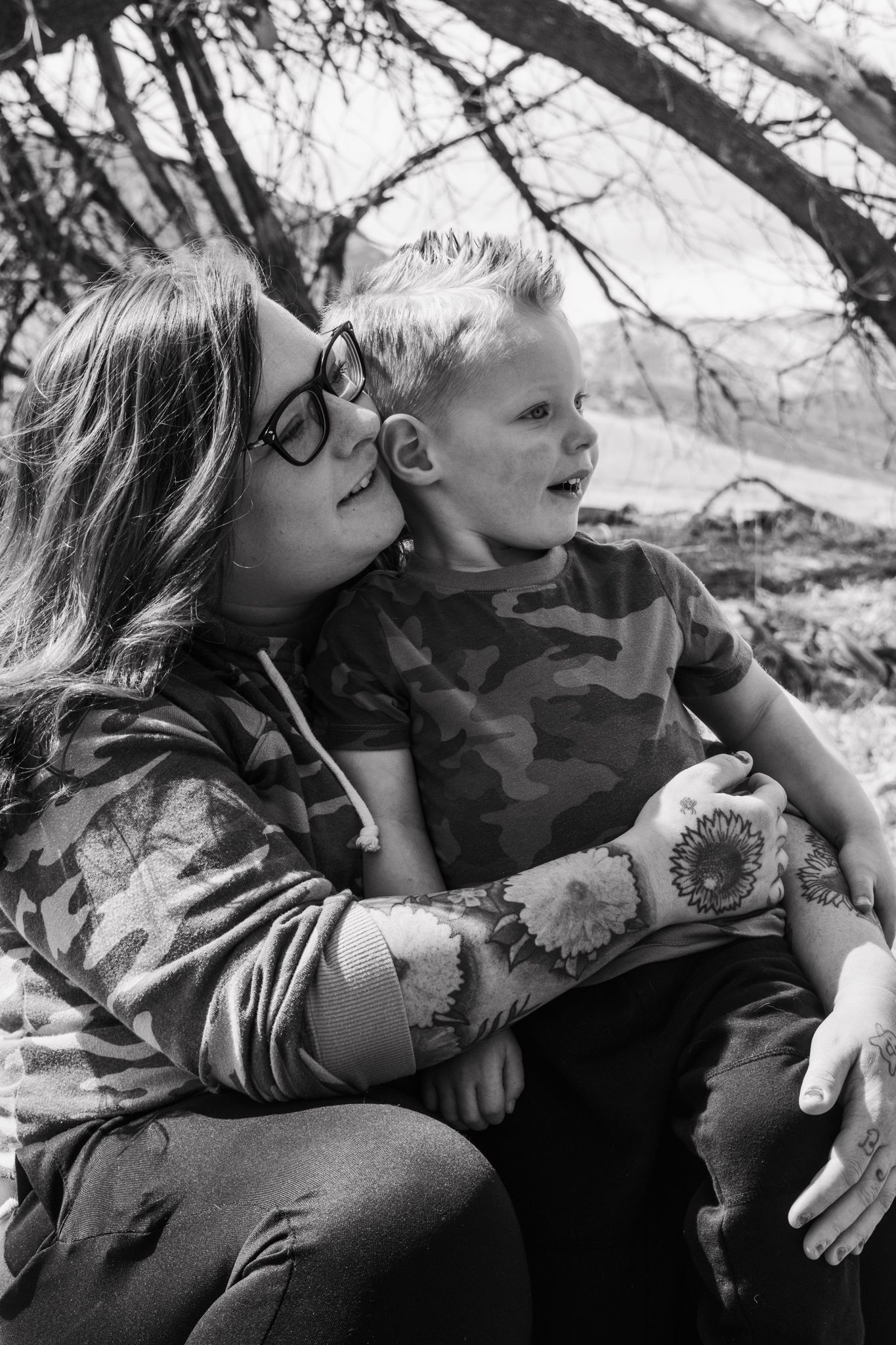 Black and white photo of a tattooed mother hugging her young son under bare trees outdoors. Family Photographer in Pflugerville, Tx