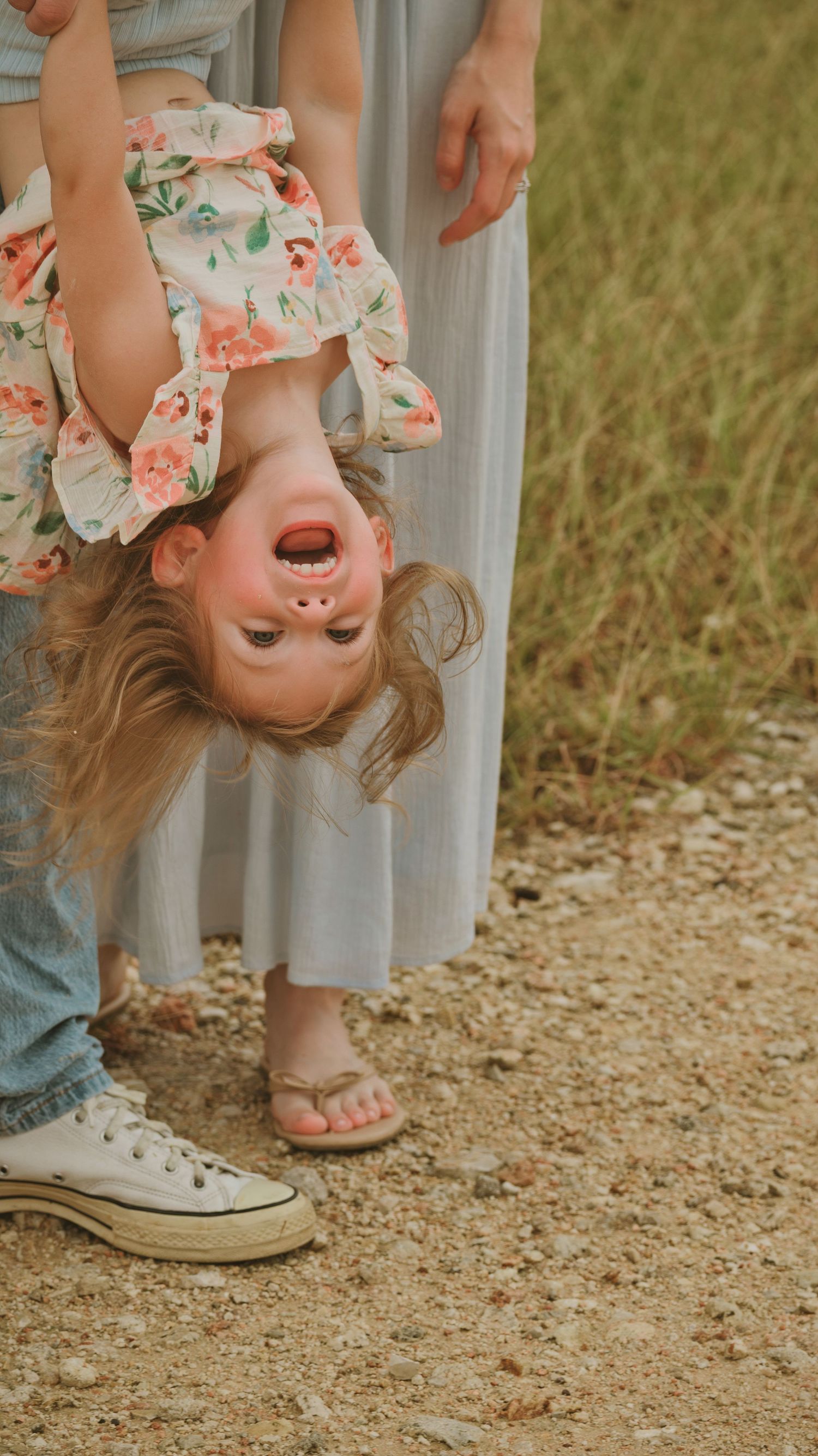 Laughing toddler girl held upside down by parents on a gravel path outdoors.