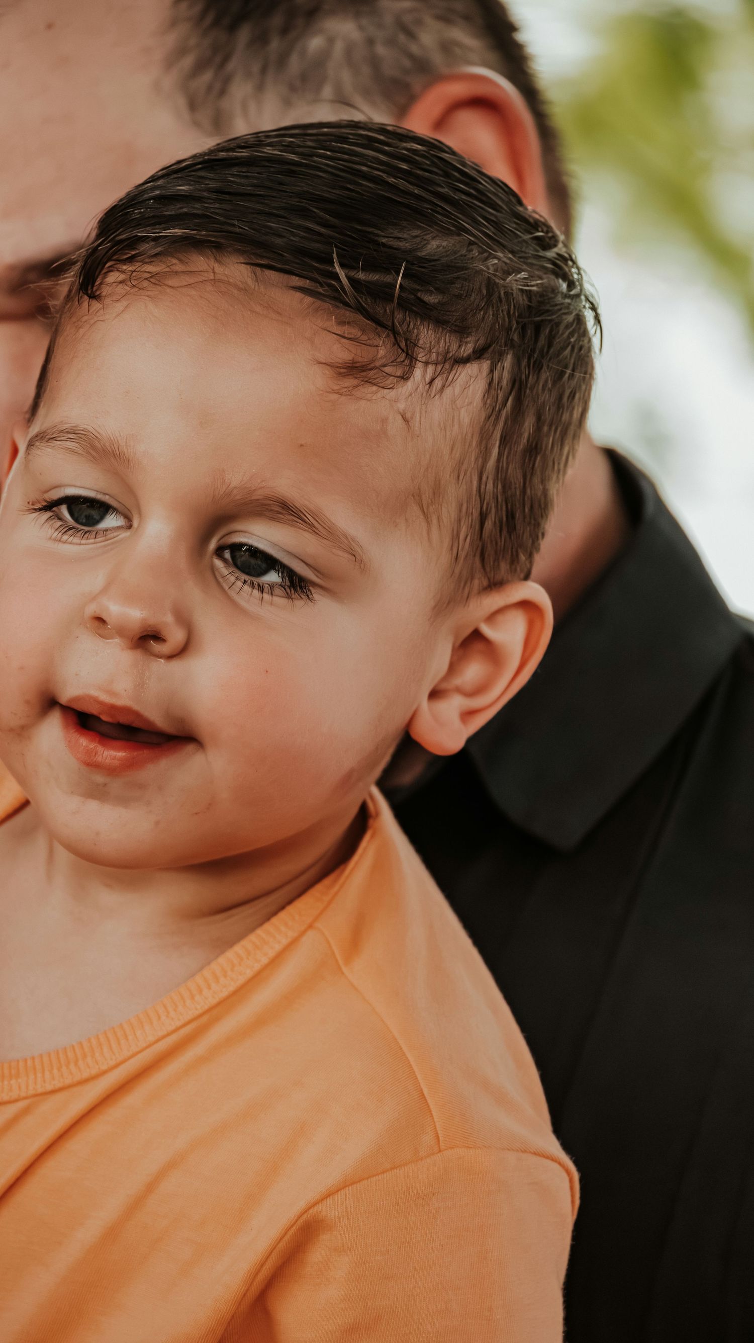Toddler boy in orange shirt smiling while being held by a man in black shirt.