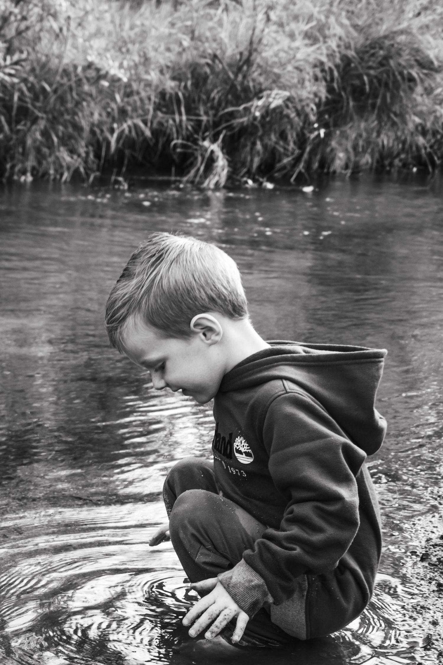 Black and white photo of a young boy crouching in a shallow river, touching the water surface with his hand.
