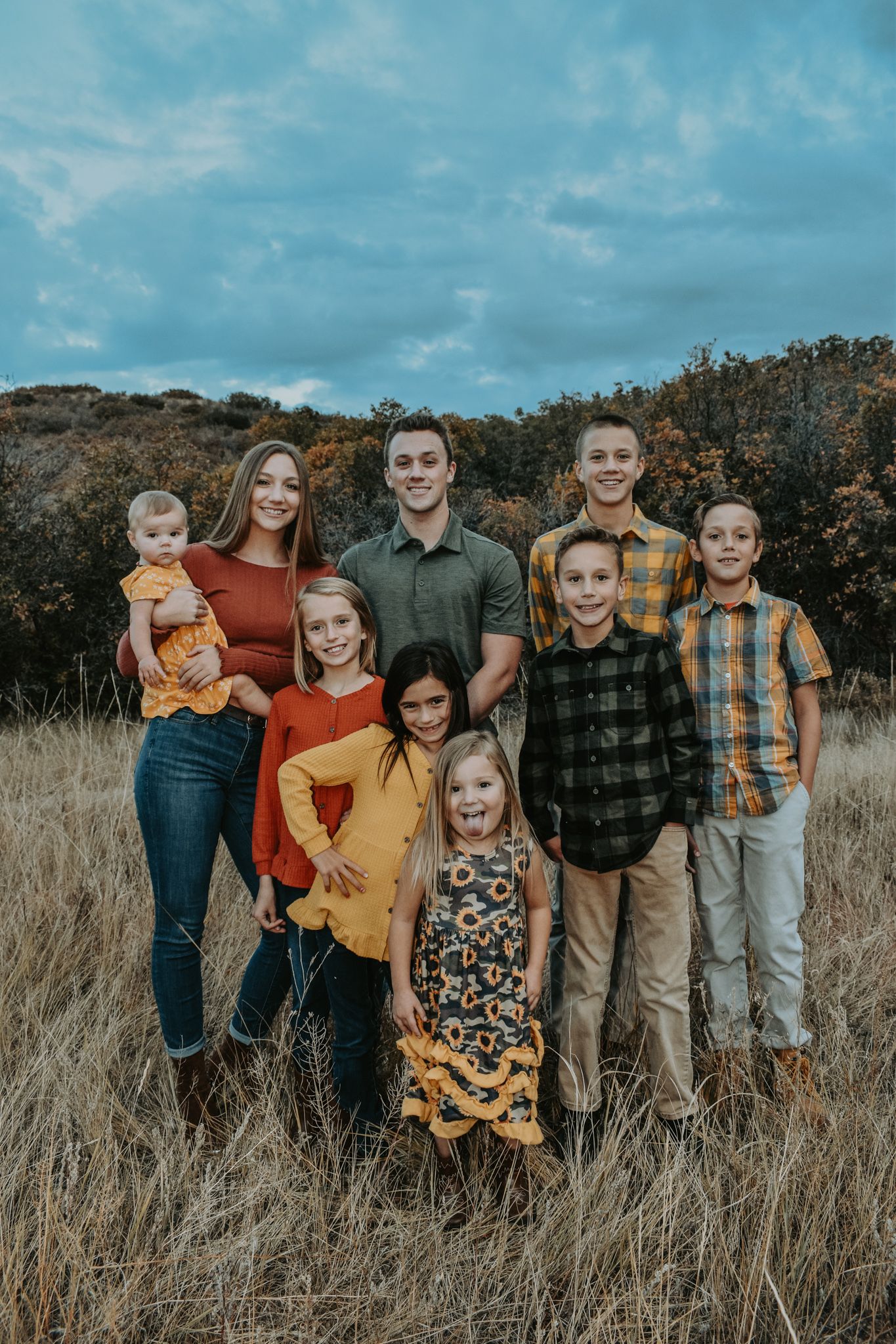 Large family group portrait of eight children posing in a golden autumn field under a cloudy sky.