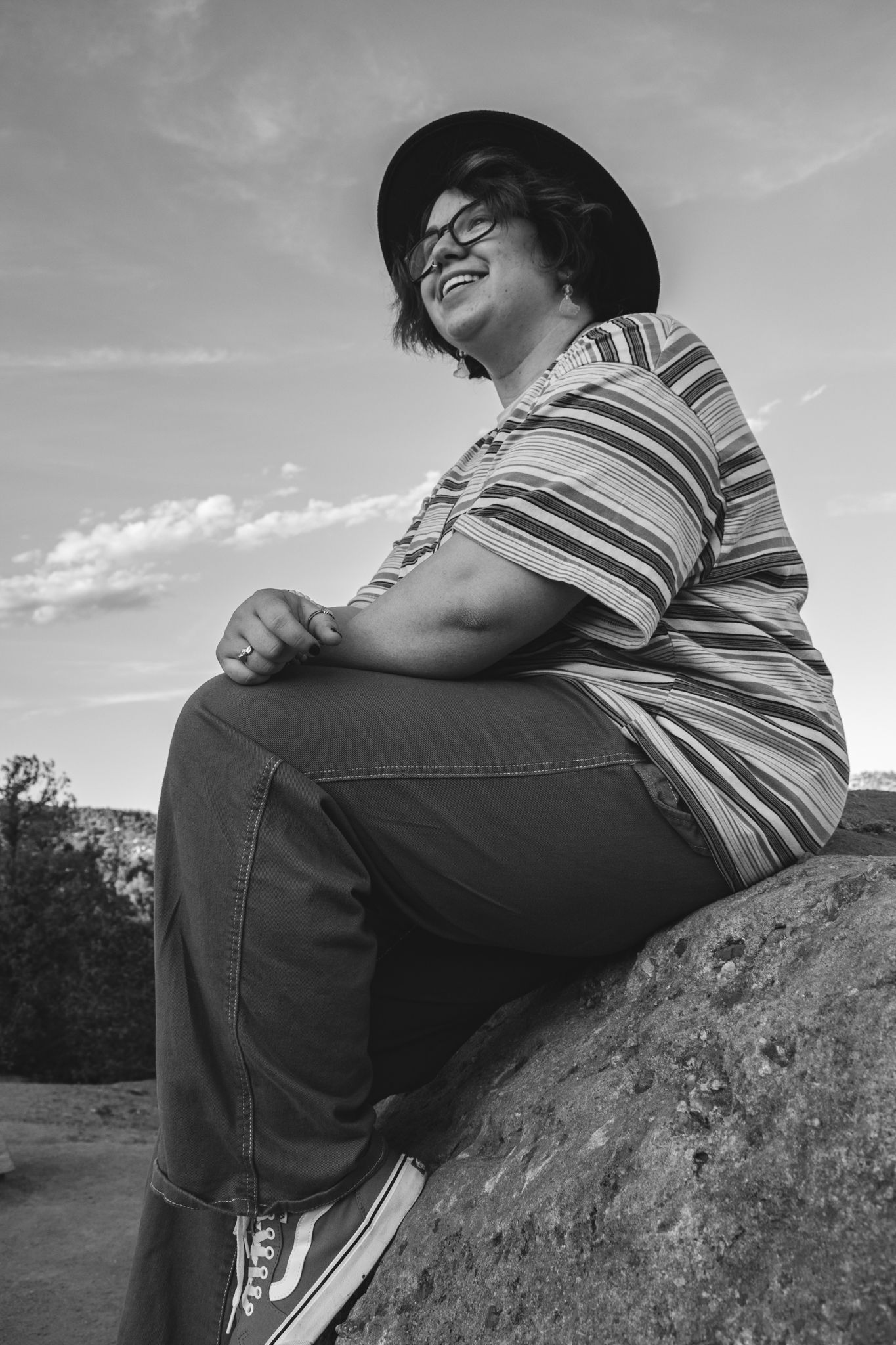 Person sitting on a rock smiling outdoors in black and white, wearing a striped shirt, hat, and glasses for senior photos.