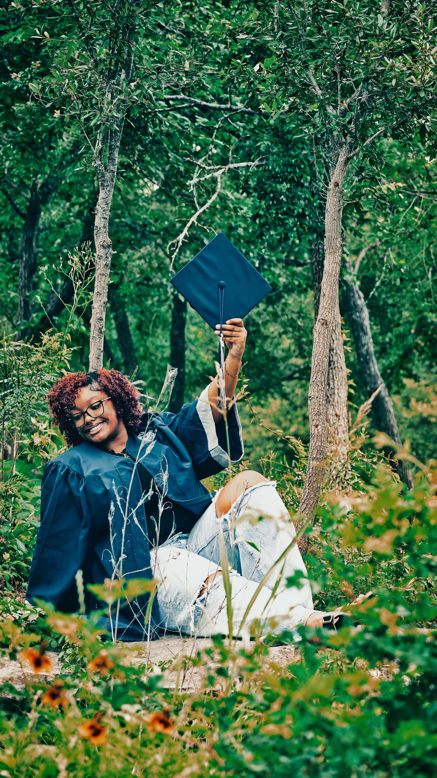 Joyful graduate in blue gown sitting in a lush green forest holding graduation cap aloft for senior photos.