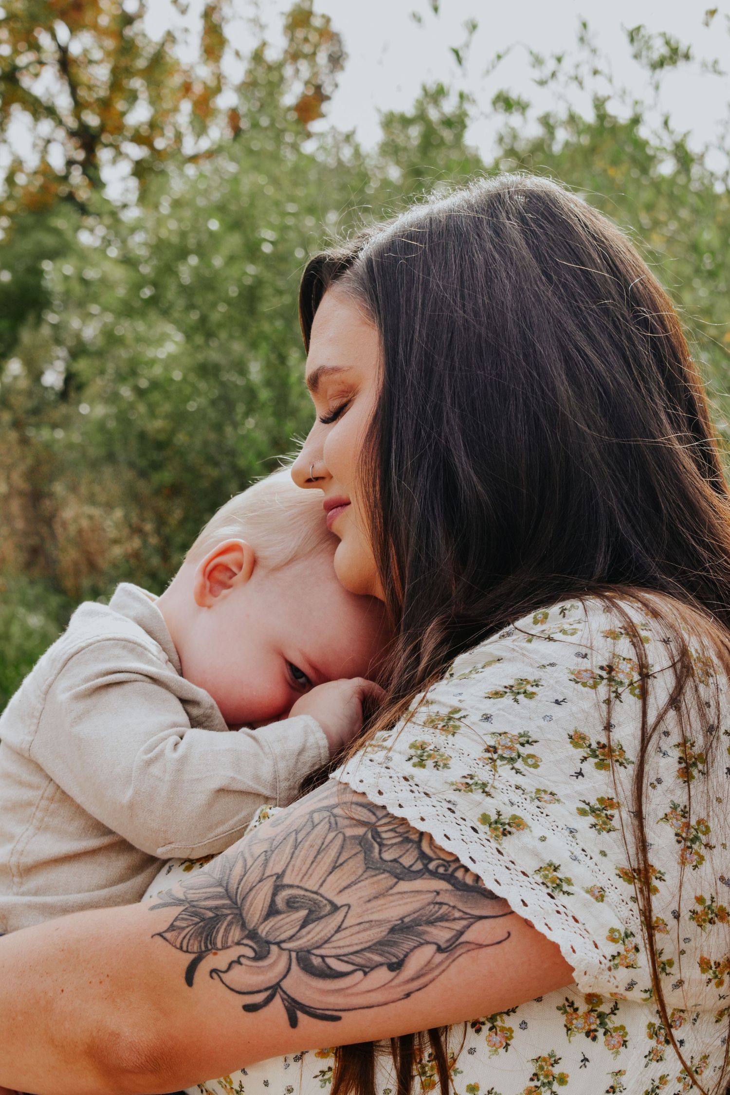 Dark-haired tattooed mother with floral tattoo tenderly nuzzling her sleeping baby outdoors.
