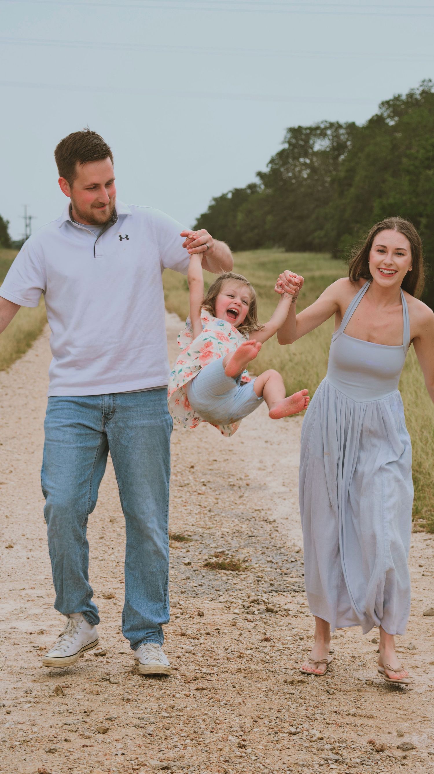 Joyful family swinging their laughing toddler by the hands on a scenic country dirt road.