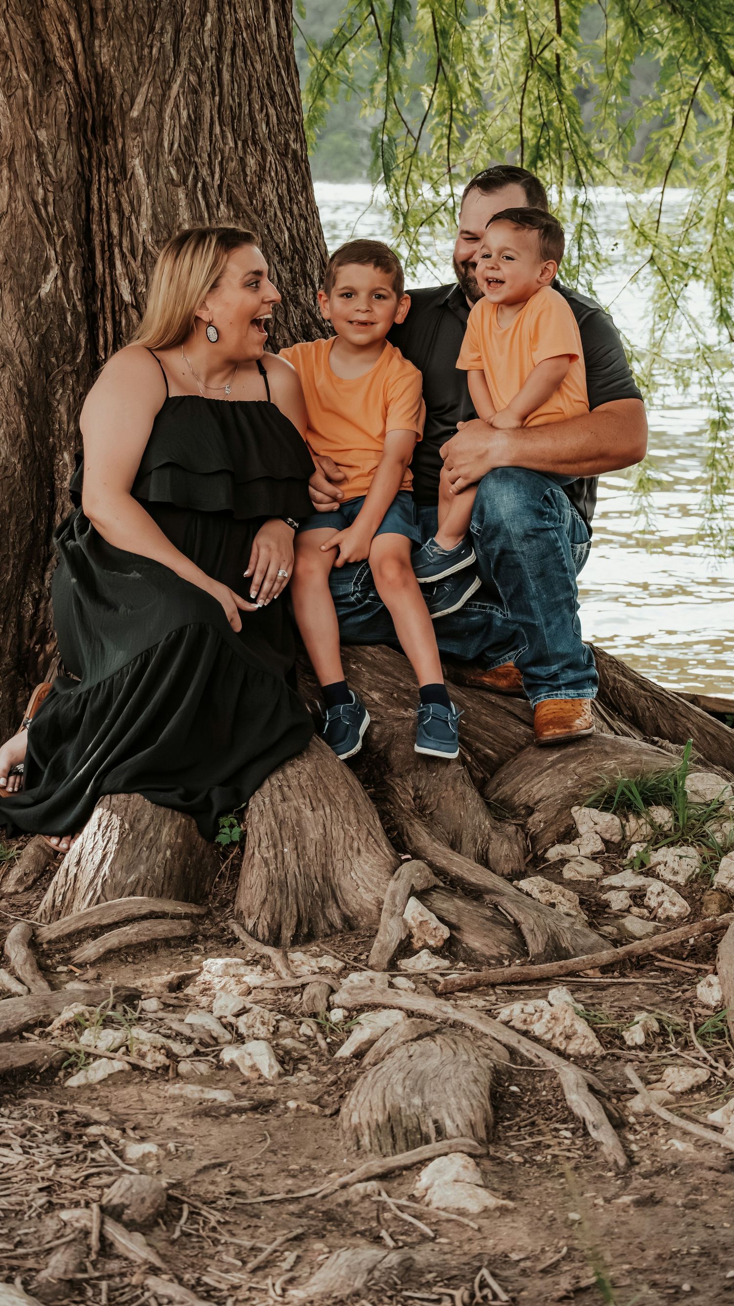 Joyful family laughing together on tree roots near a lake, two boys in orange shirts.