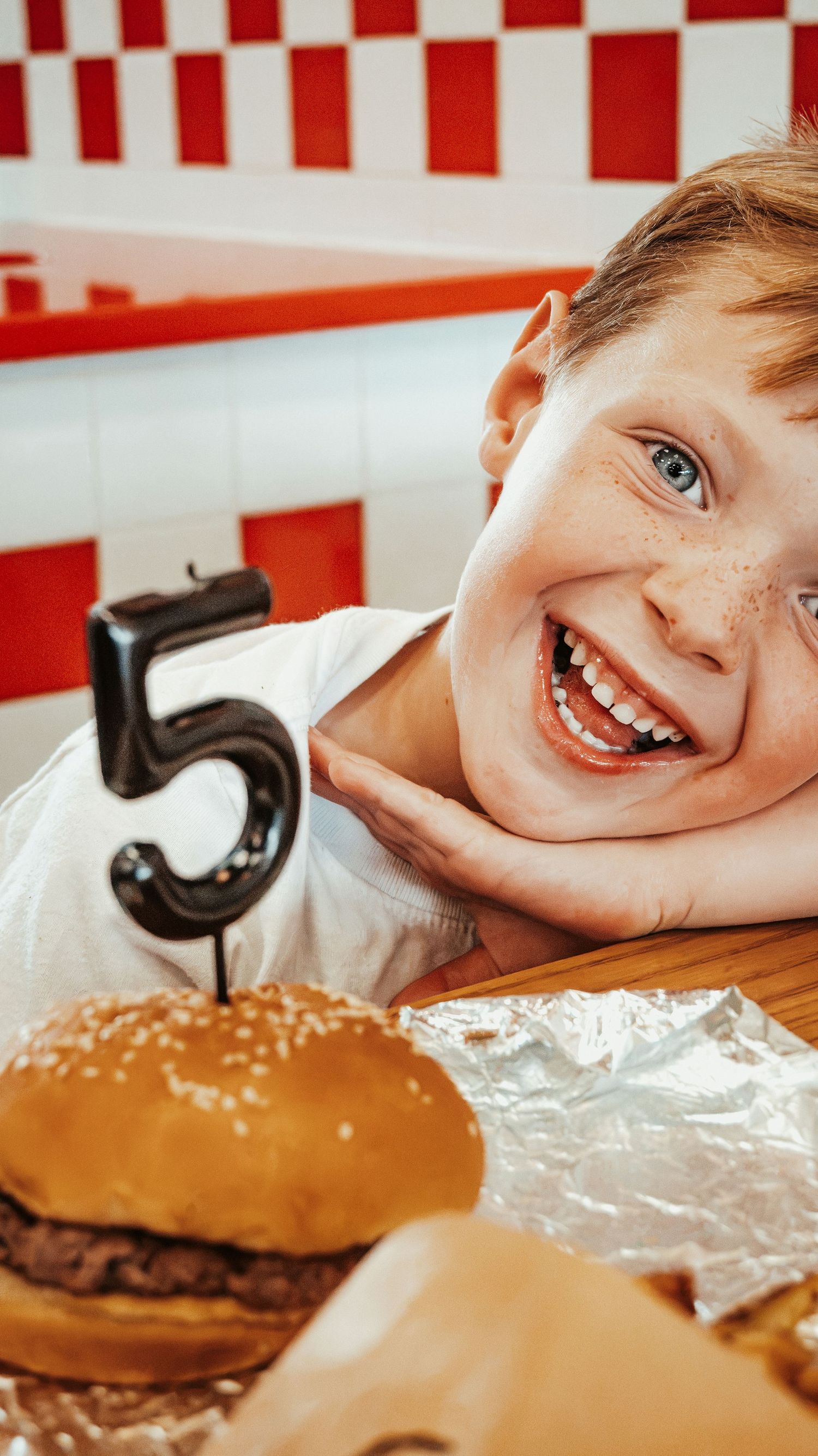 Smiling redhead child at Five Guys restaurant with a burger and number 5 candle. Family Photographer in Pflugerville, Tx