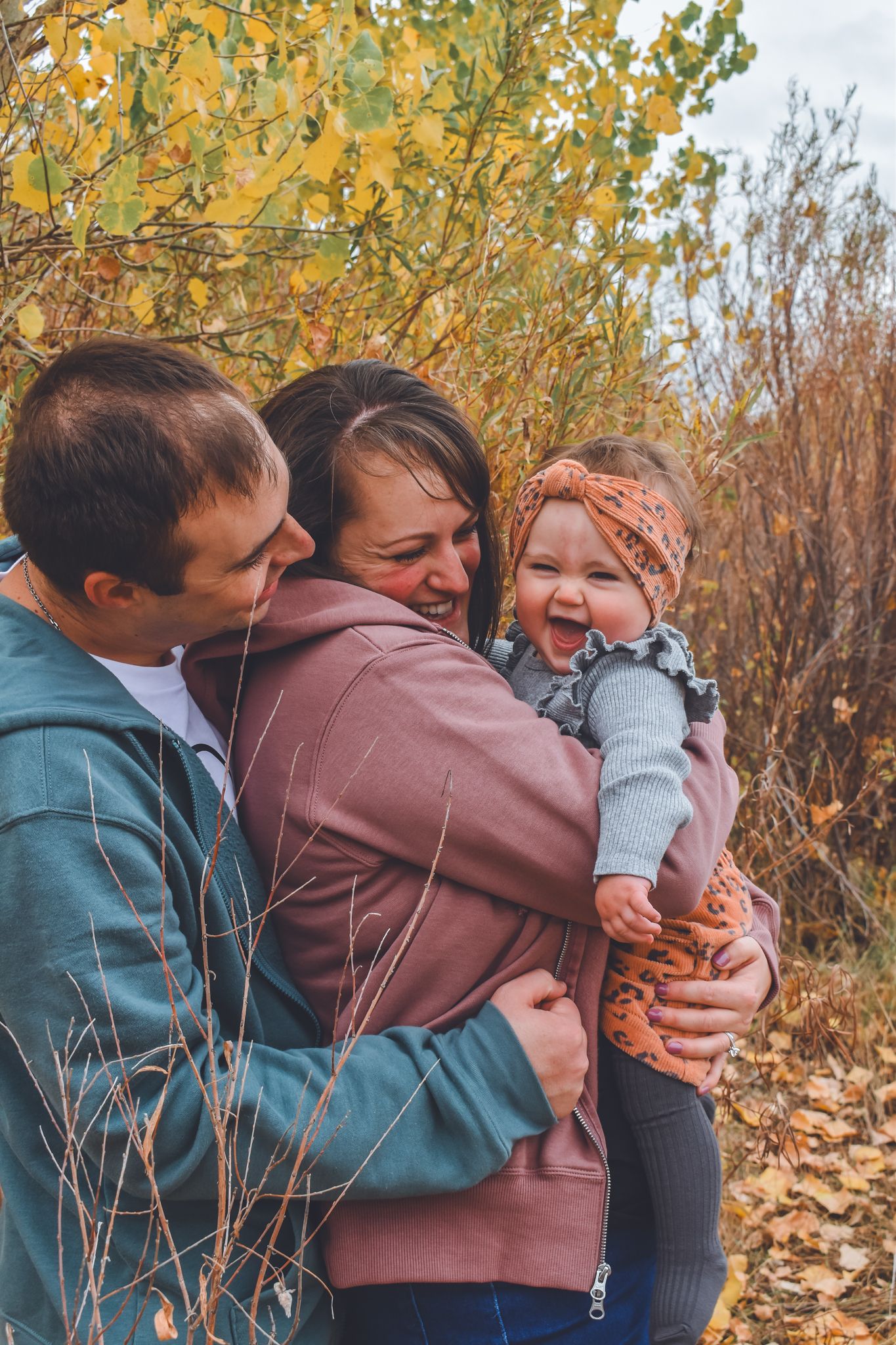 A happy family of three shares a joyful embrace outdoors surrounded by vibrant yellow autumn foliage. Family Photographer in Pflugerville,