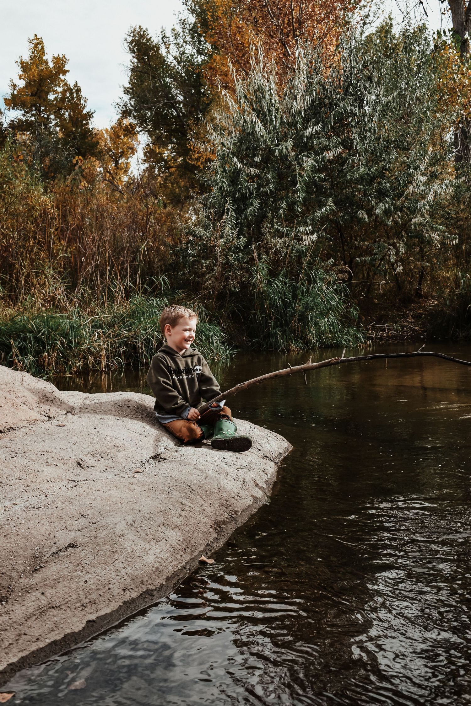 Young boy sitting on a rock by a calm river fishing with a stick rod in autumn nature setting.