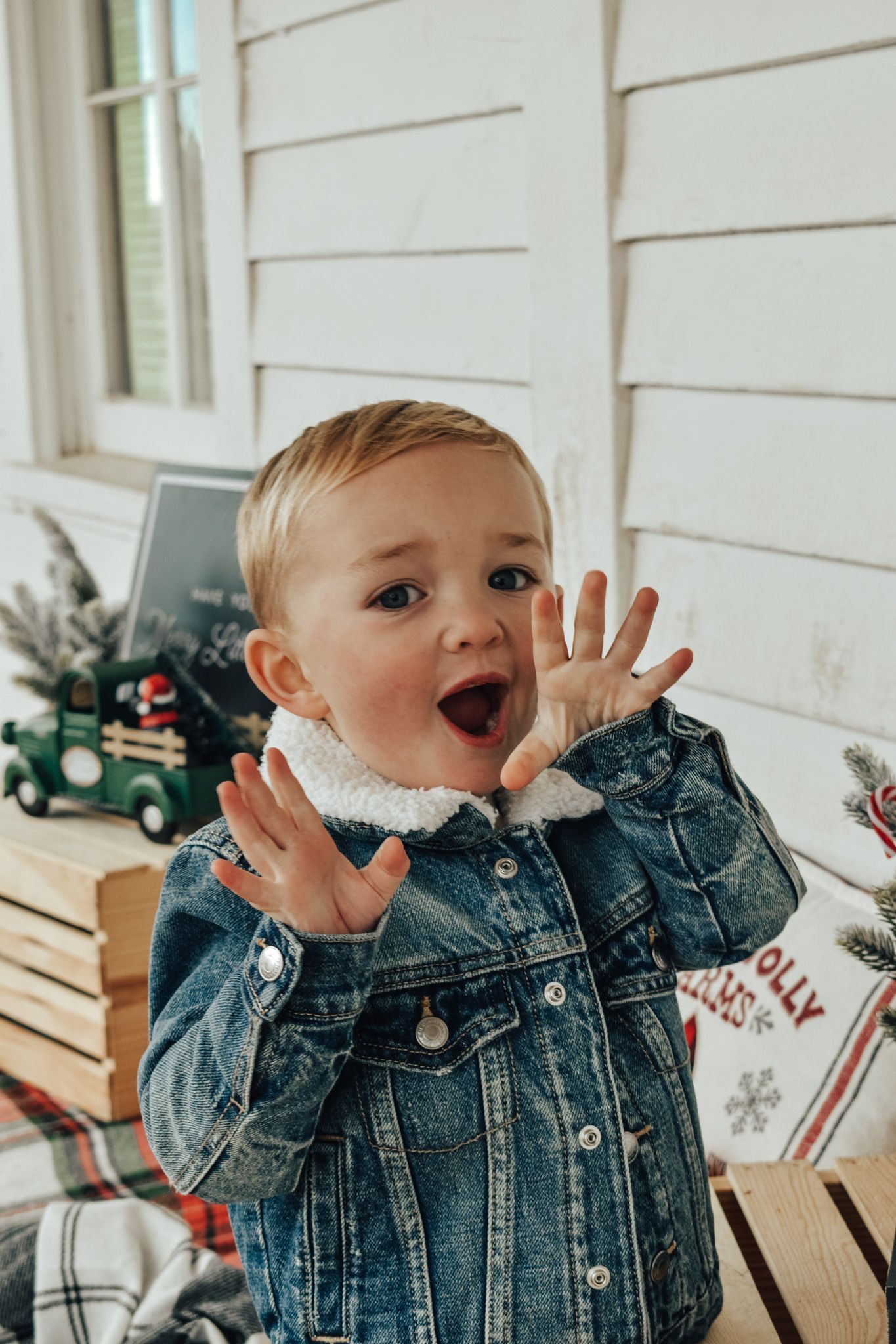 Surprised toddler in denim jacket with hands raised near Christmas decorations.